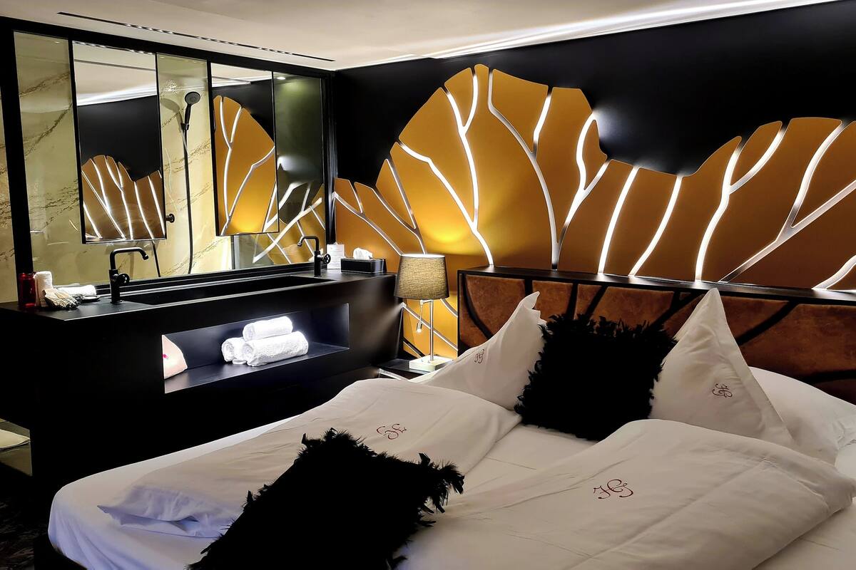 A hotel room with a modern design, featuring a large bed with white linens, a black headboard with a unique leaf-like pattern, a black vanity with a mirror, and a wall-mounted light fixture.