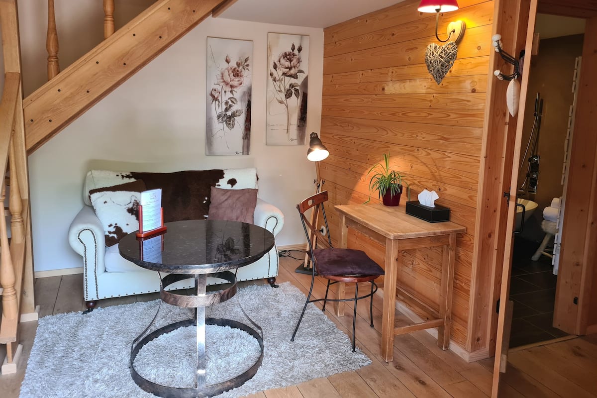 A cozy room with a wooden staircase, a white sofa with a patterned throw, a round table, a small wooden side table with a plant, and a wall-mounted lamp.