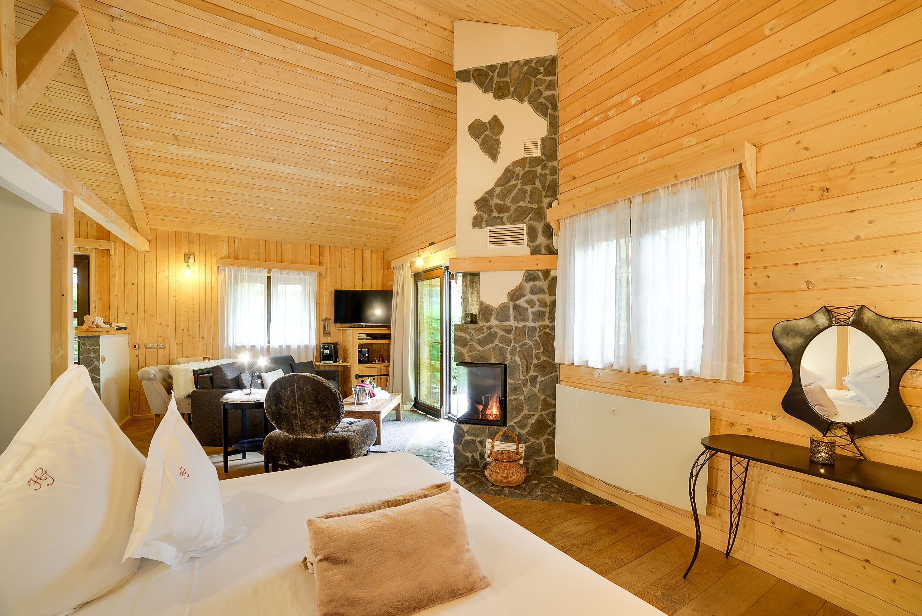 A cozy log cabin room with a stone fireplace, wooden walls, a bed with pillows, a desk, and a television.