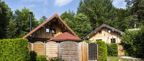 A wooden cabin with a sloped roof, surrounded by dense greenery and a gravel path.