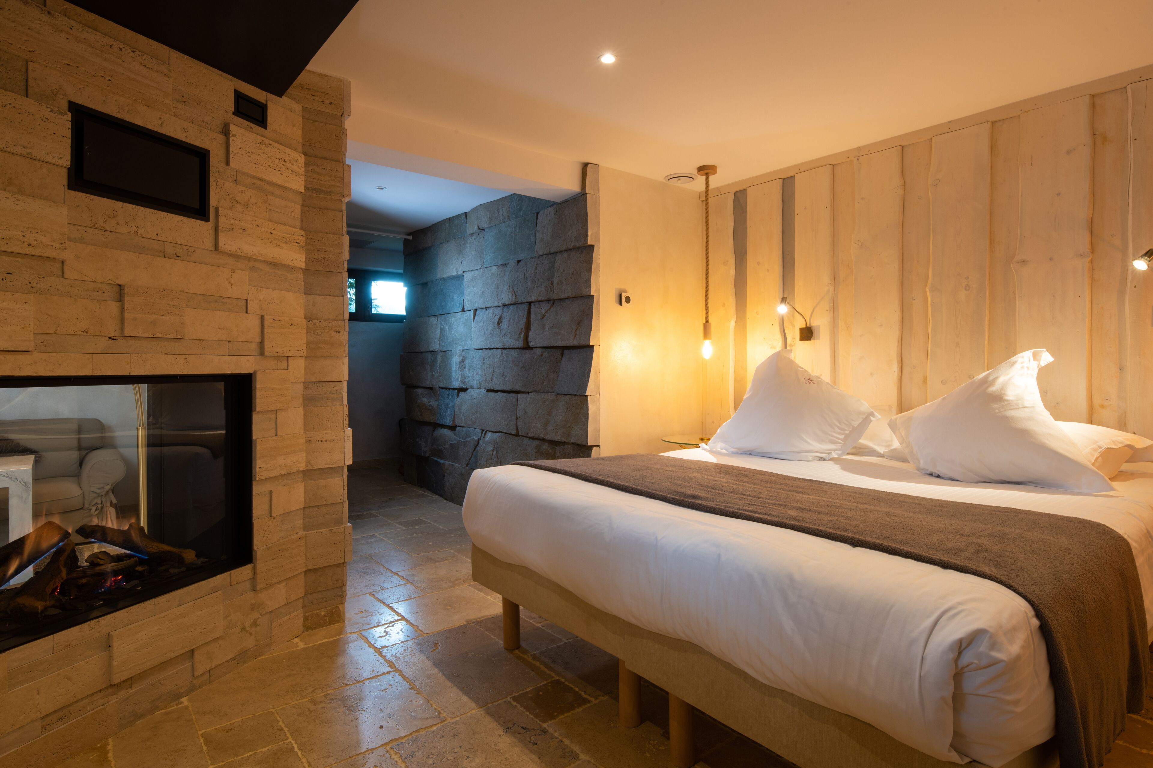 A modern bedroom with a stone wall, a fireplace, a large bed with white bedding, and a wooden headboard.