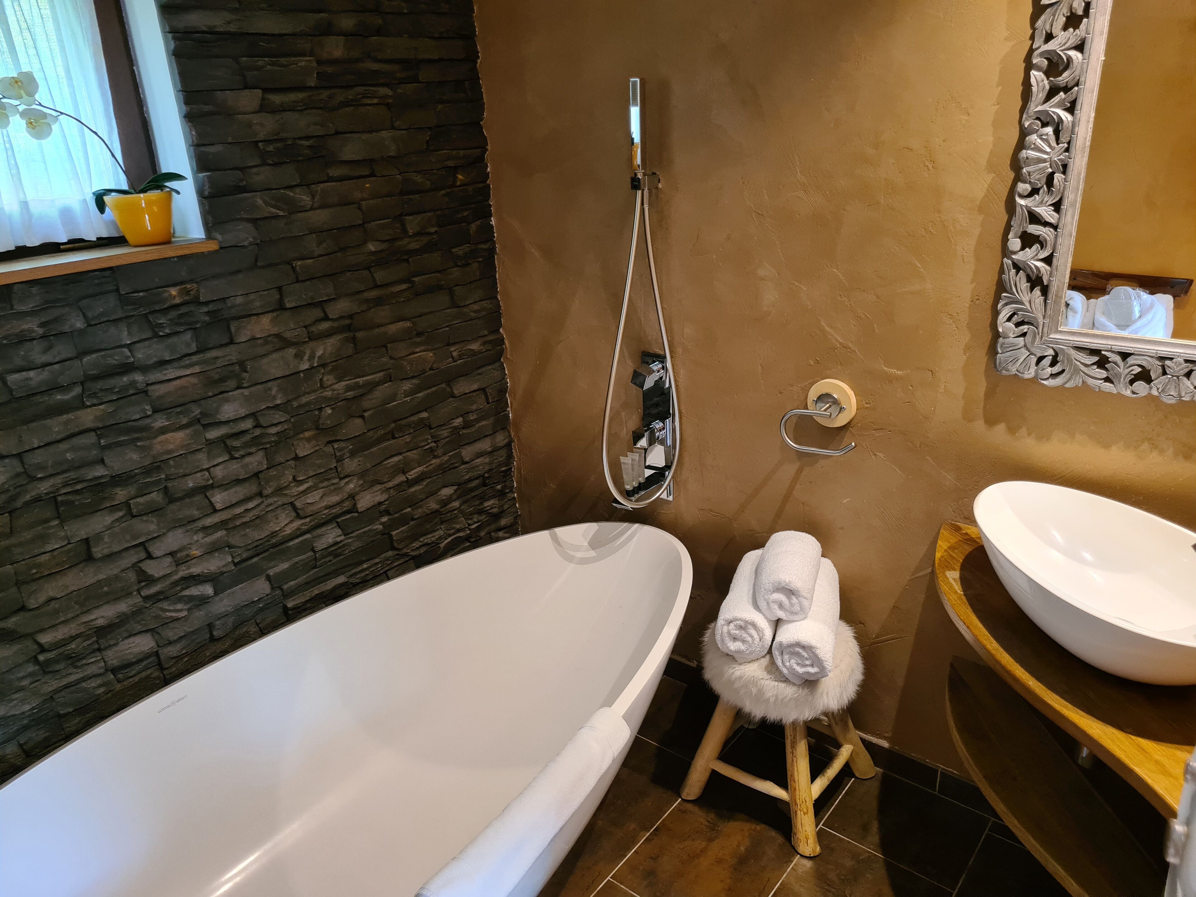 A bathroom with a stone wall, a white bathtub, a wooden stool with towels, a white sink, and a mirror with a floral frame.