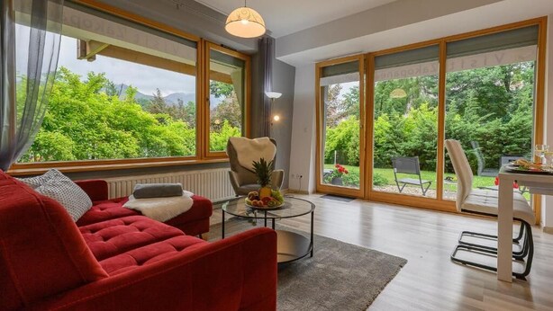 Ein modernes Wohnzimmer mit einem roten Sofa, einem Glastisch mit Obst und großen Fenstern mit Blick auf Grünflächen.