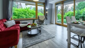 Ein modernes Wohnzimmer mit einem roten Sofa, einem runden Couchtisch und einem Essbereich mit weißem Tisch und Stühlen. Große Fenster bieten Blick auf Grünflächen außerhalb.