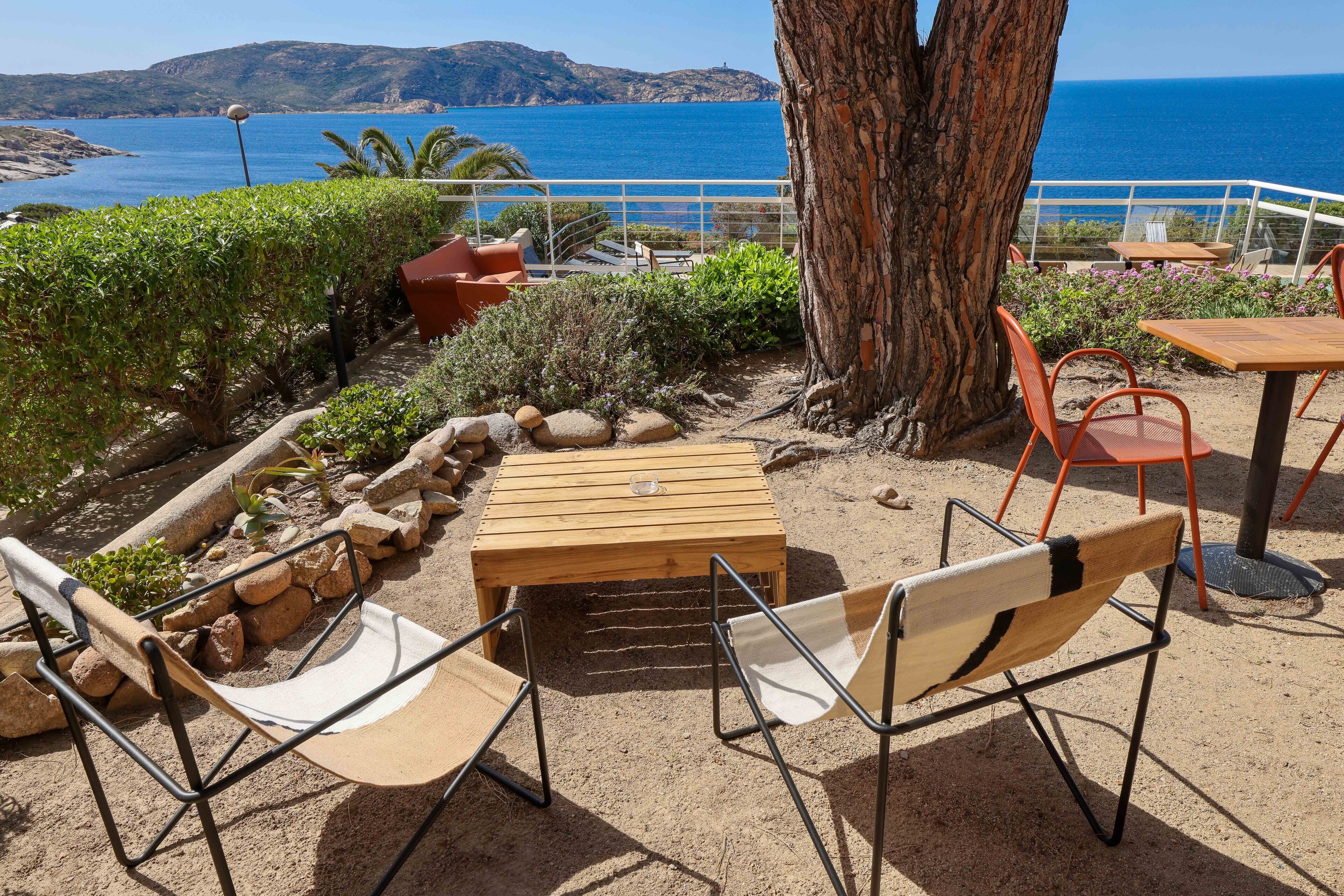An outdoor seating area with a wooden table and chairs, a tree, and a view of the sea.
