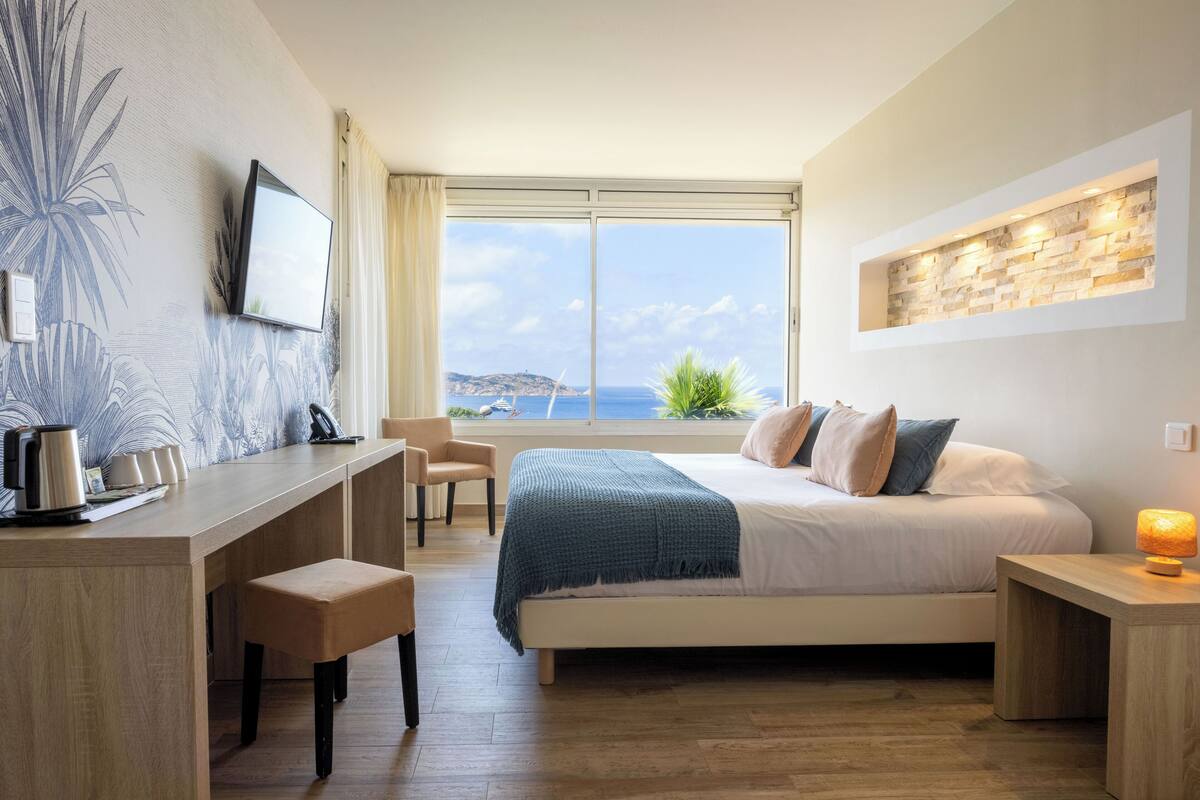 Quarto com cama, mesa de cabeceira, escrivaninha e cadeira. Há uma TV na parede, uma janela com vista para o mar e um objeto decorativo na parede.