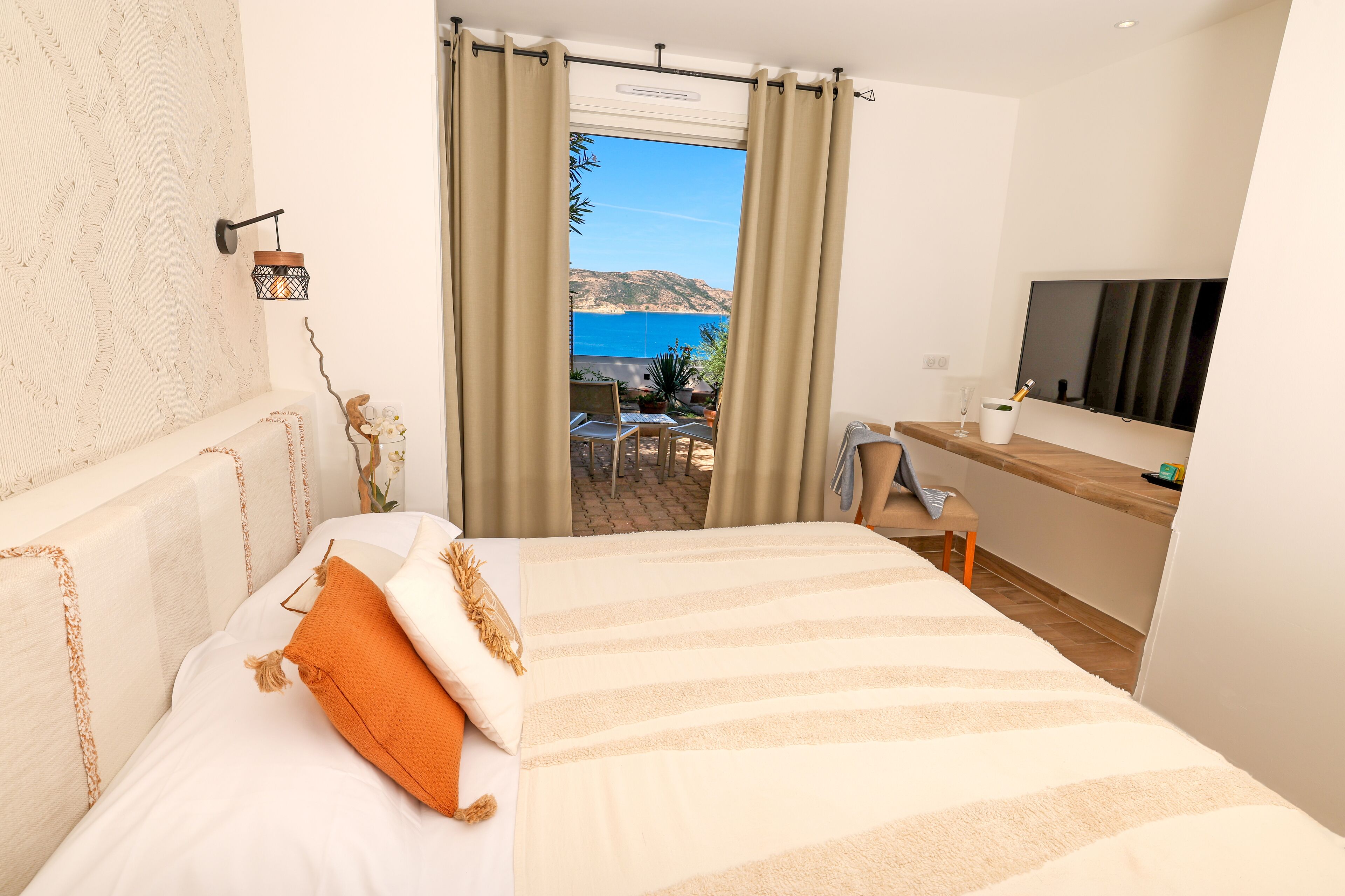 A bedroom with a bed, a desk, a TV, and a view of the sea.