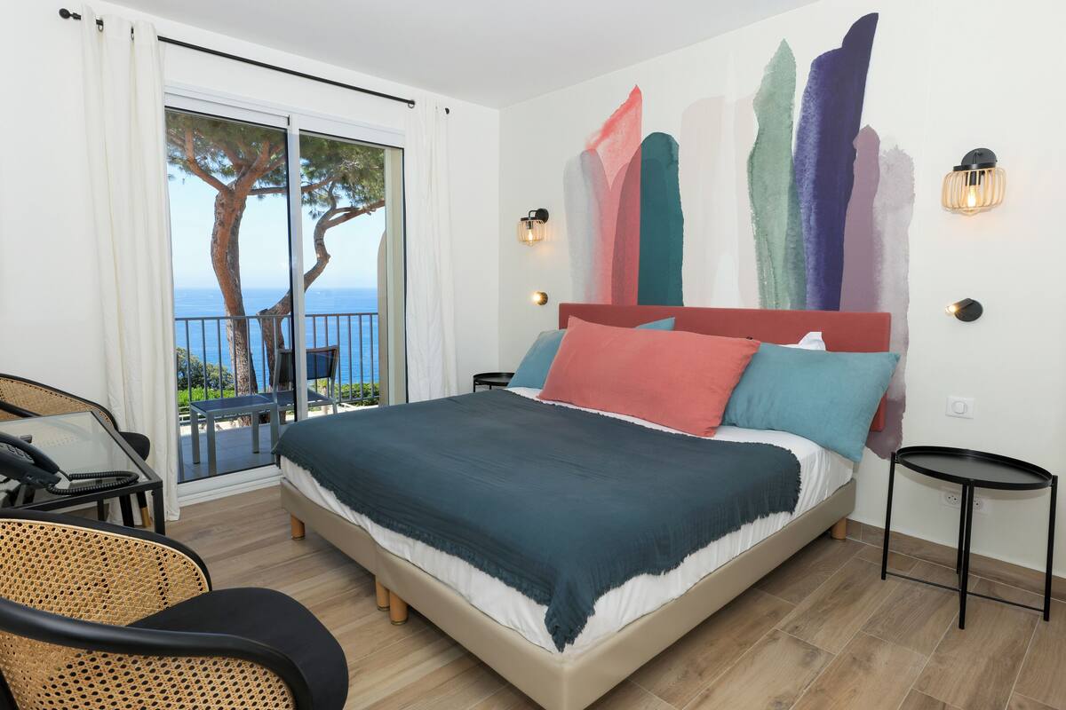Quarto com cama, uma mesinha, uma cadeira e vista para o exterior.