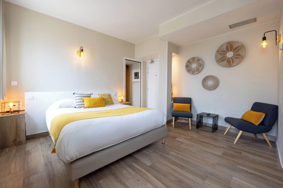 Quarto com cama grande, mesas de cabeceira, uma cadeira, arte decorativa na parede e porta que dá para outro cômodo.