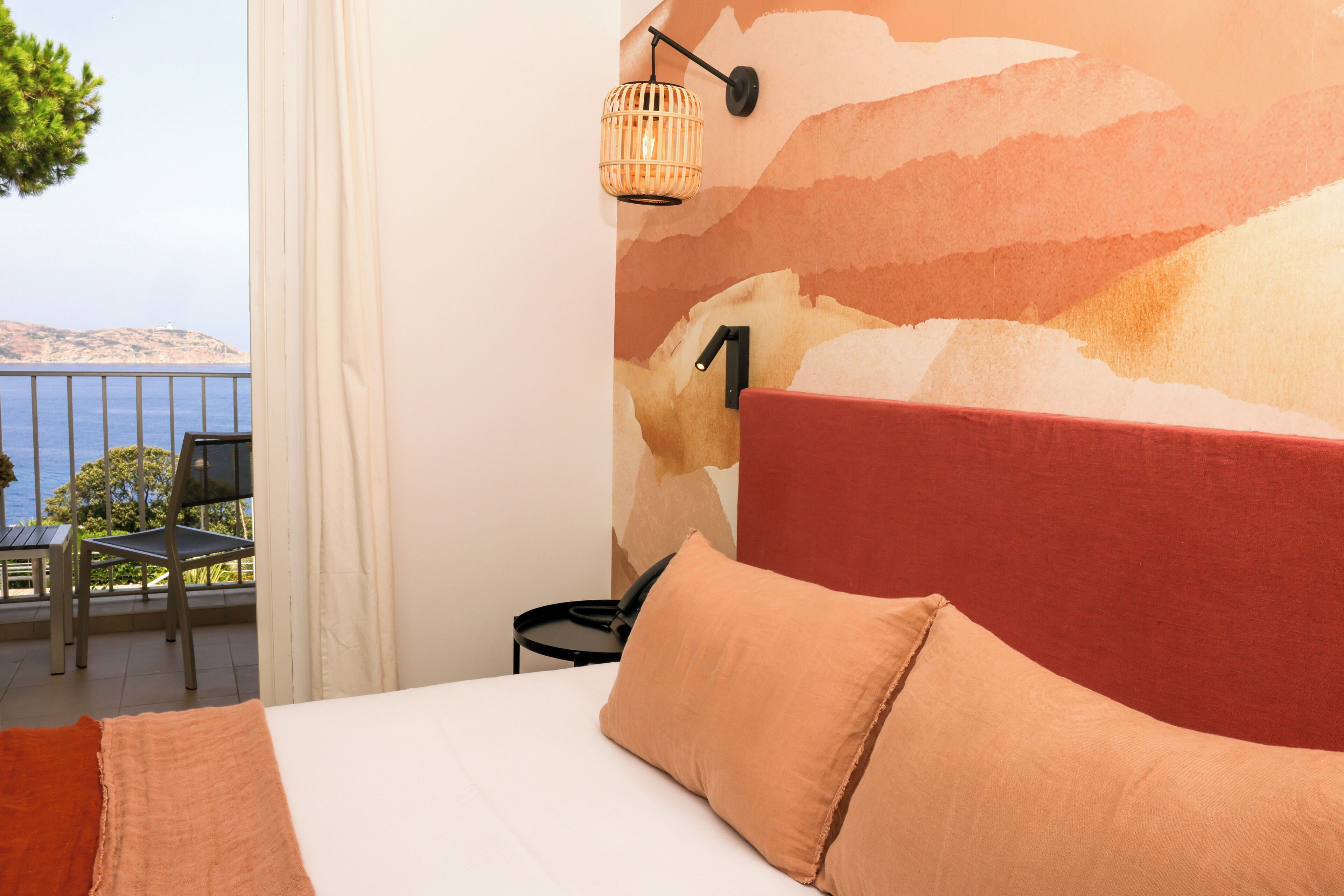 A hotel room with a bed, a red headboard, a wall-mounted lamp, and a view of the sea.