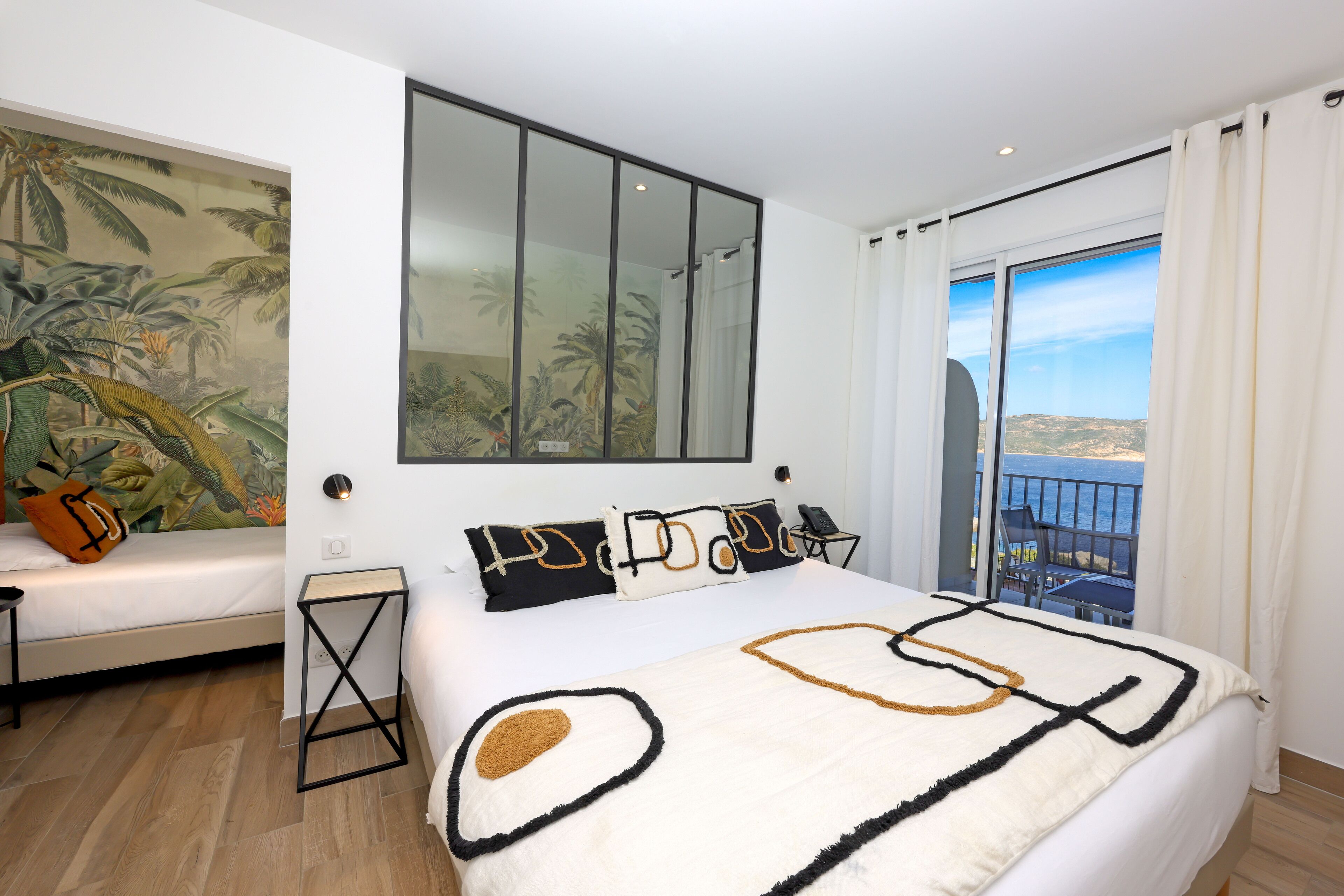 A bedroom with a large bed, a nightstand, a wall-mounted TV, and a balcony with a view of the sea.