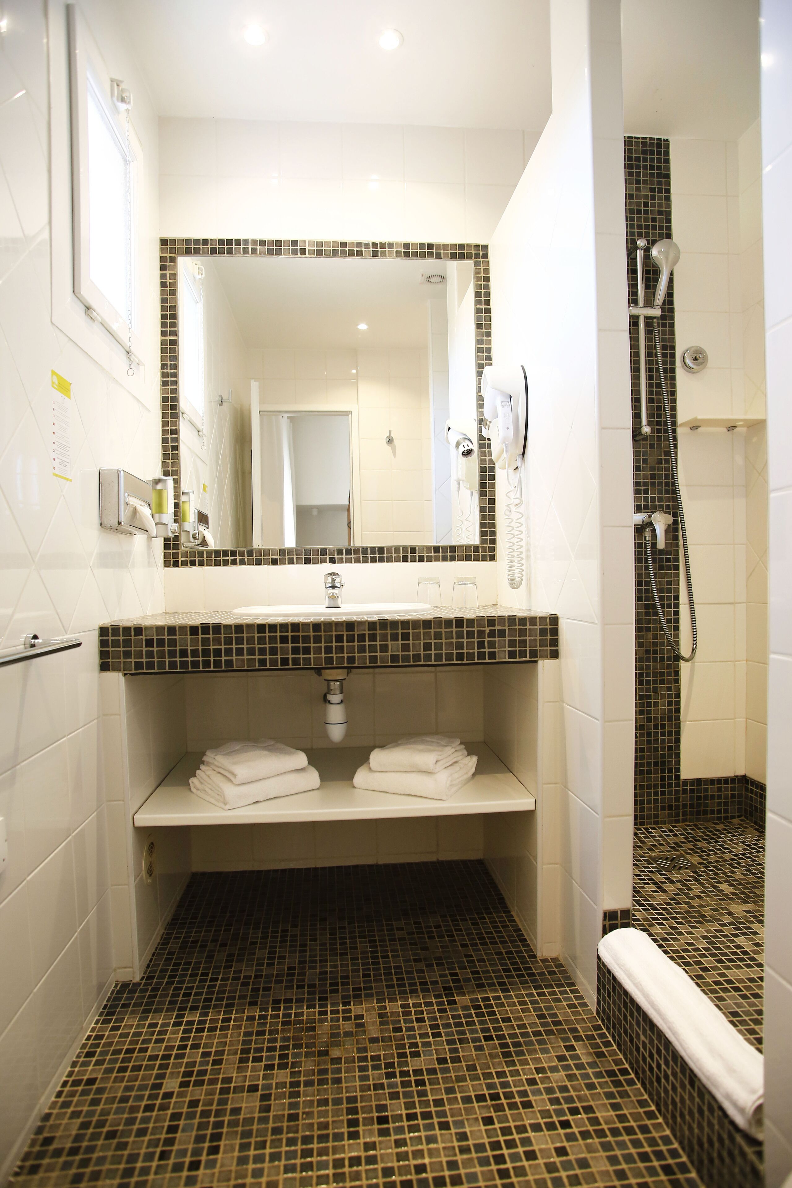 Une salle de bain avec un sol carrelé, un lavabo avec un miroir au-dessus, un porte-serviettes et un espace douche.
