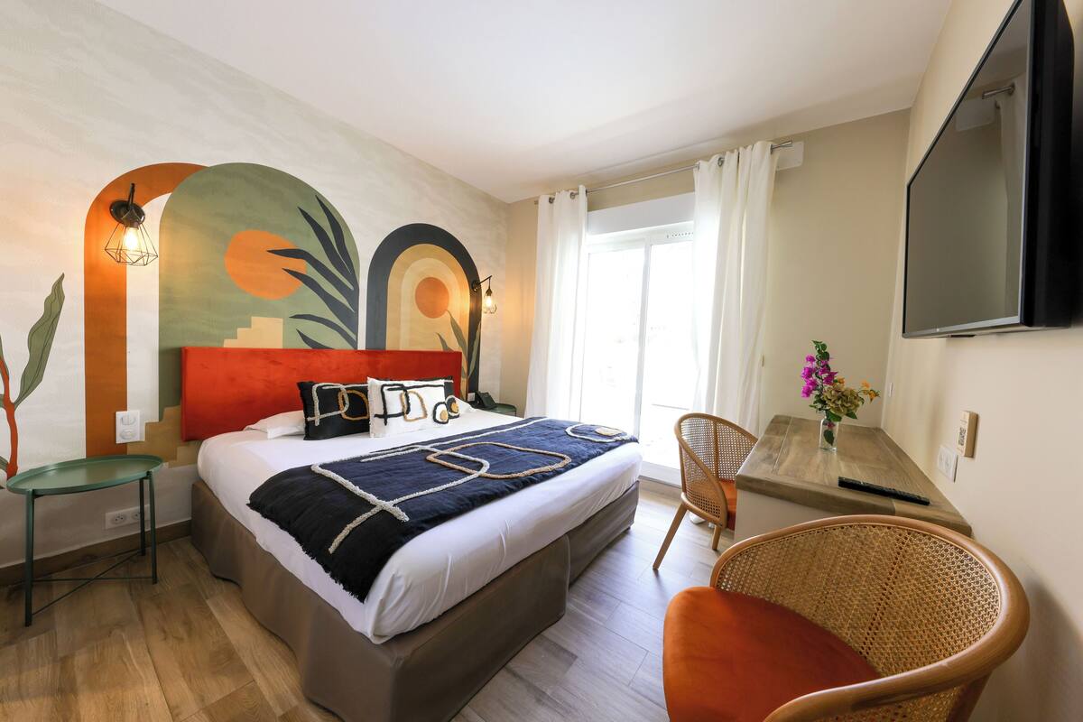 Quarto de hotel com cama, escrivaninha, cadeira, televisão e janela com cortinas.
