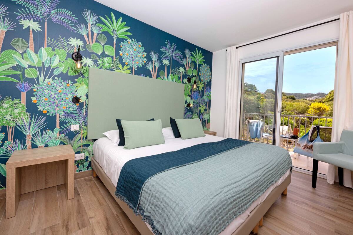 Quarto com uma cama grande, uma mesa de cabeceira, vista para o exterior por uma porta de correr de vidro e um mural de plantas tropicais na parede.