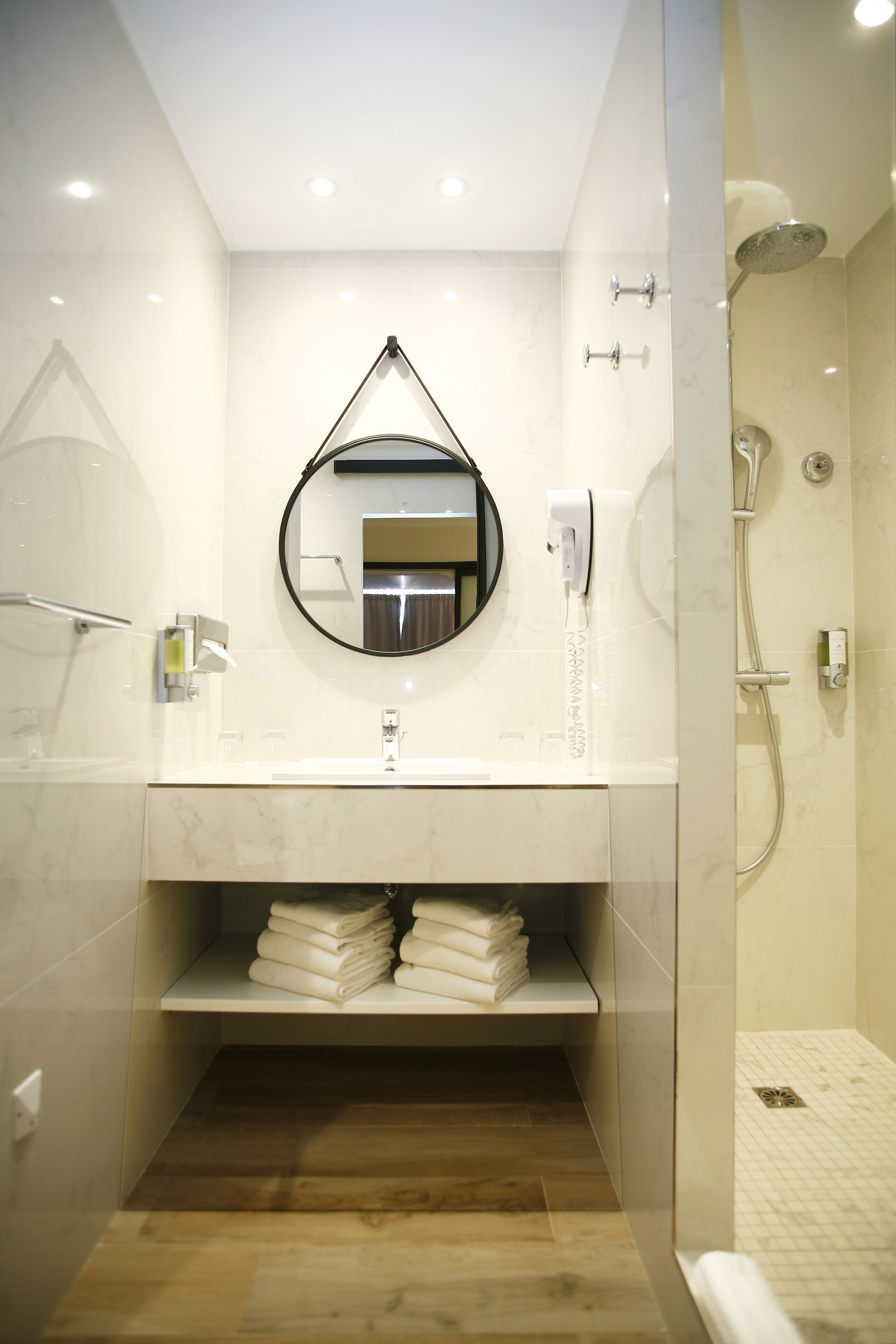 Un baño moderno con tocador de mármol, un espejo redondo, un estante empotrado con toallas dobladas y una zona de ducha con cabezal de ducha manual.