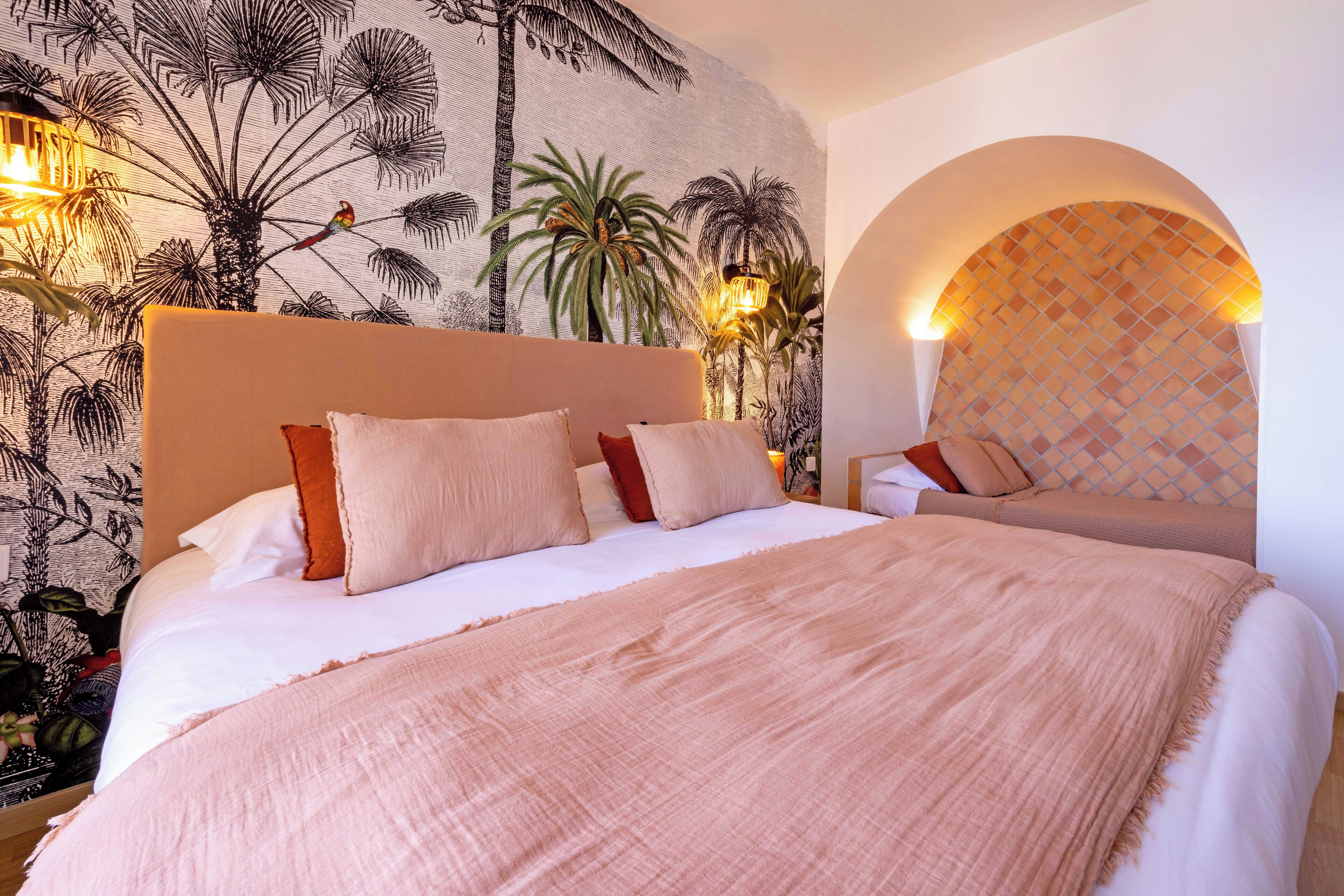 A neatly made bed with a pink blanket and multiple pillows, a wall with a tropical mural, and a small table with a lamp.