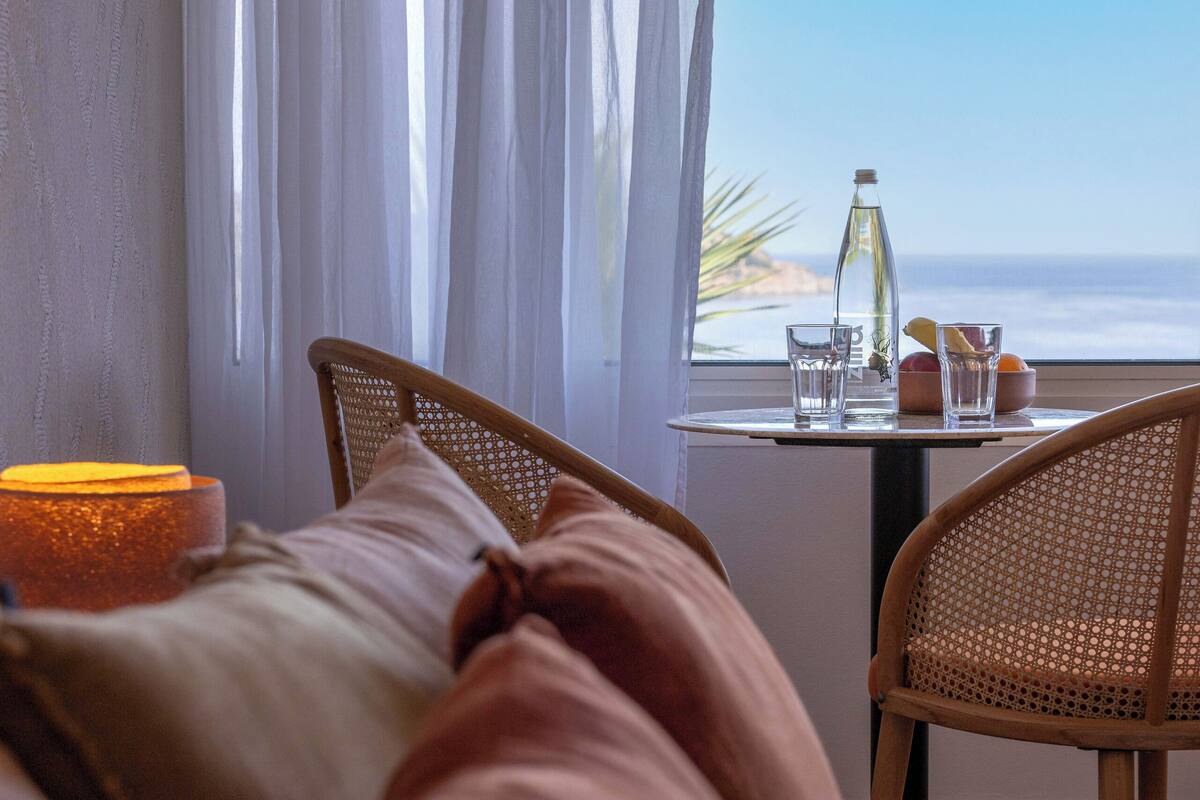 Quarto de hotel com uma cama, uma mesa com garrafa e copos, e vista para o oceano.