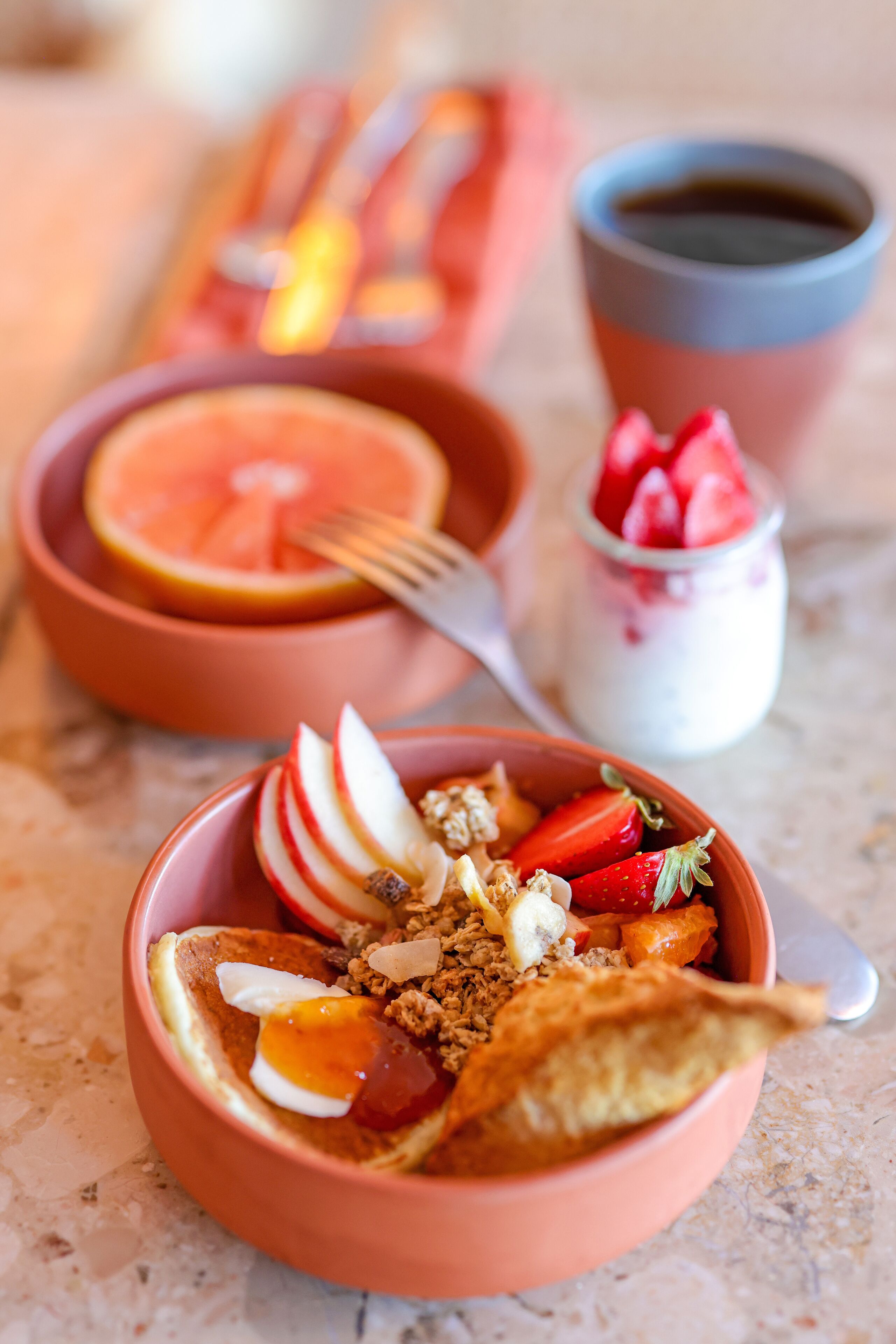 Une table mise pour le petit-déjeuner avec un bol de céréales garni de fraises, une tranche d’orange et un petit pot de yaourt.