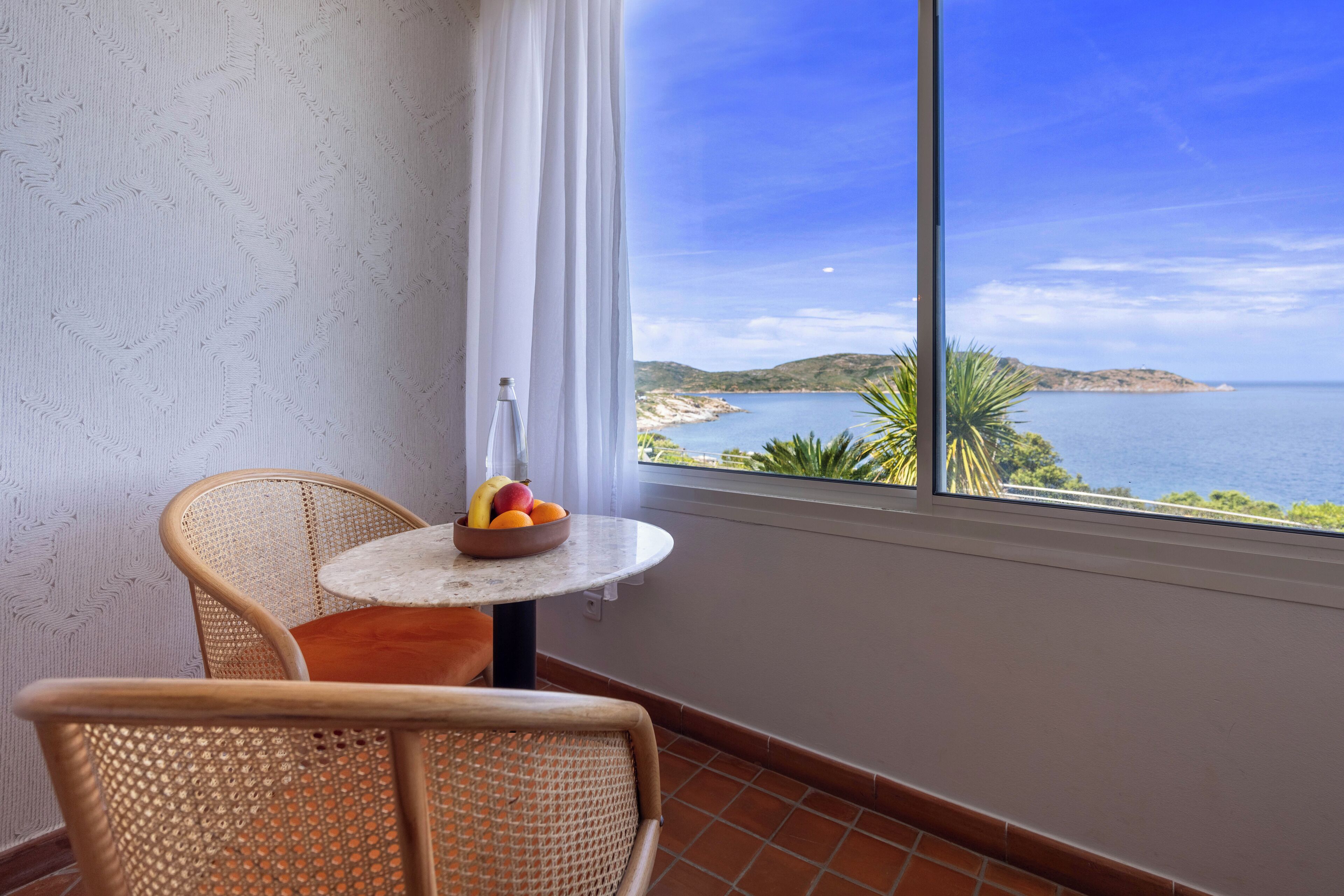 A balcony with wicker chairs, a small round table with a fruit bowl, and a view of the sea and hills.