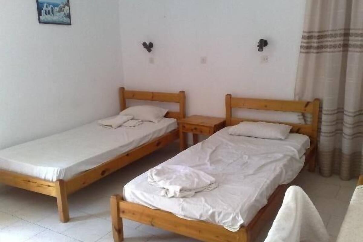 Two wooden single beds with white linens in a room with white walls and a tiled floor.