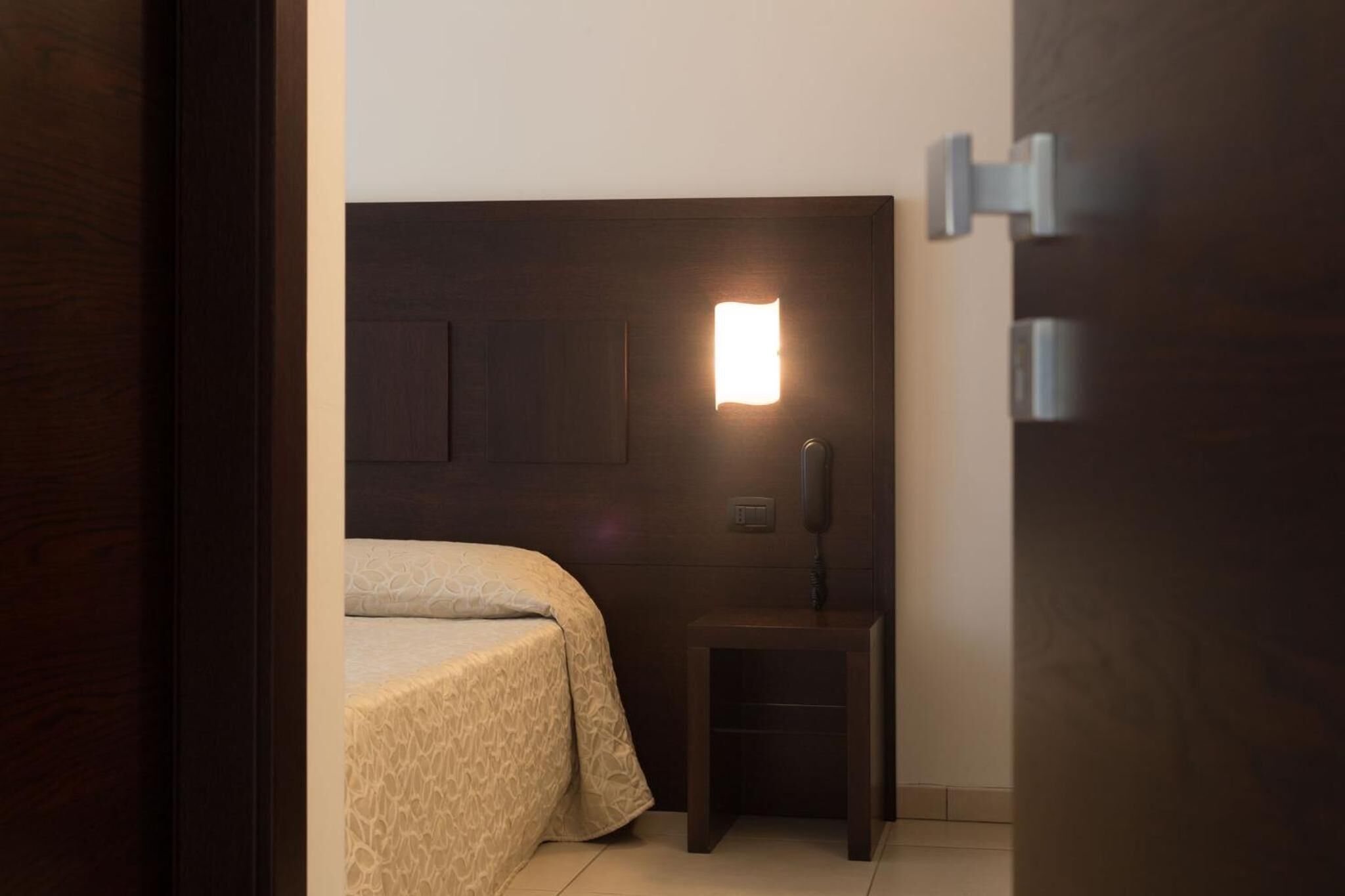 Una habitación pequeña, con poca luz, que tiene una cama individual, una mesita de noche y una lámpara en la pared.