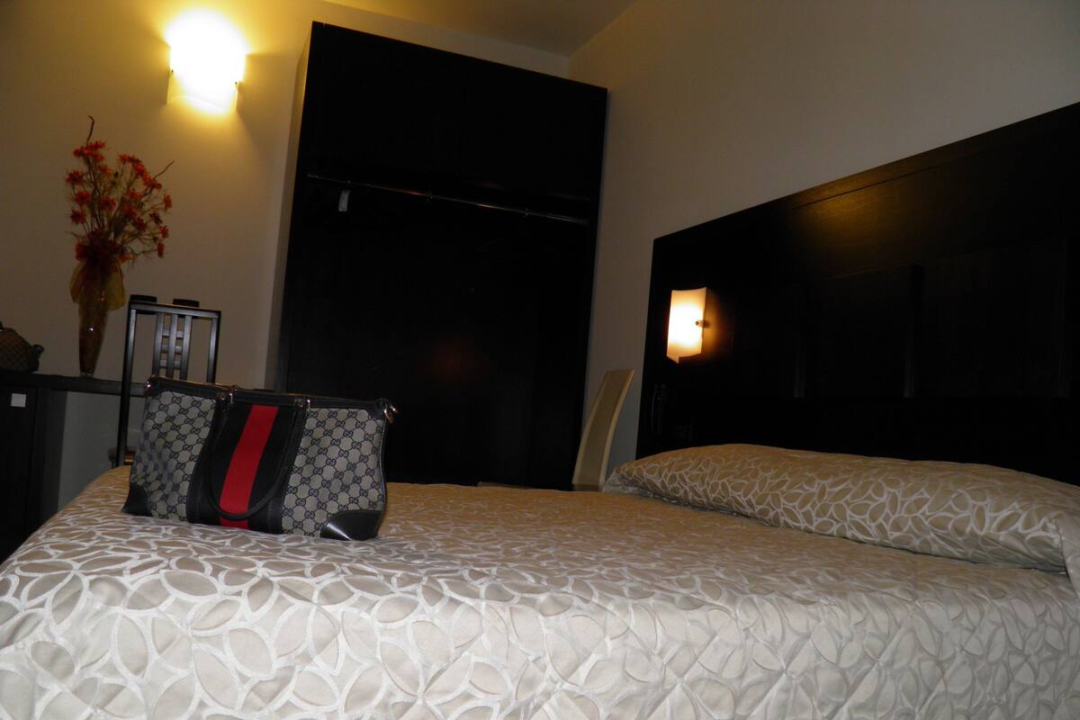 A hotel room with a bed, a chair, a handbag, and a vase with flowers.