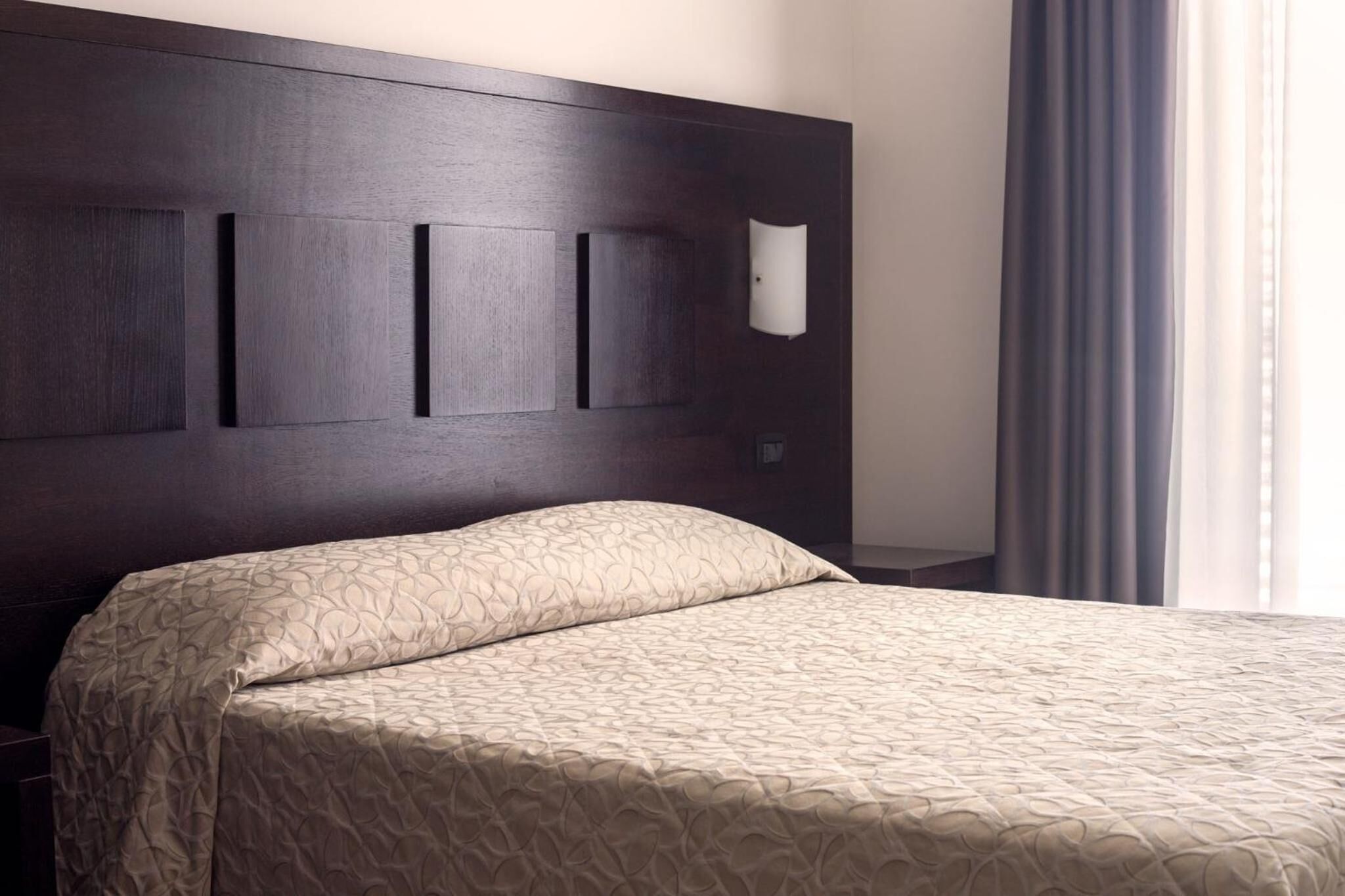 Ein Hotelzimmer mit dunkelholzvertäfelter Kopfteile, einer beigen Bettdecke mit Muster und einer weißen Wandleuchte.