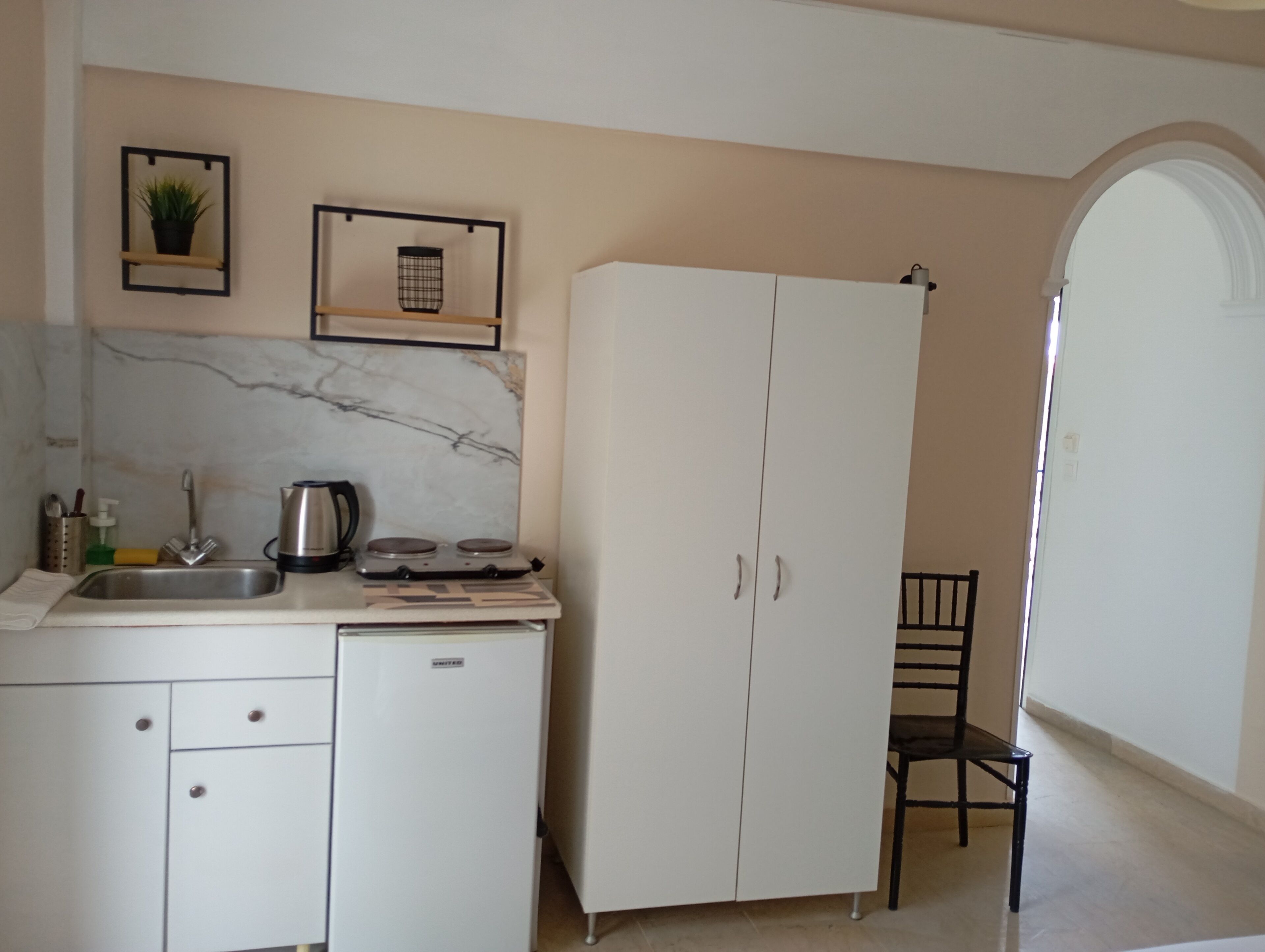 A kitchen with a sink, stove, and cabinets. There is a chair and a door leading to another room.