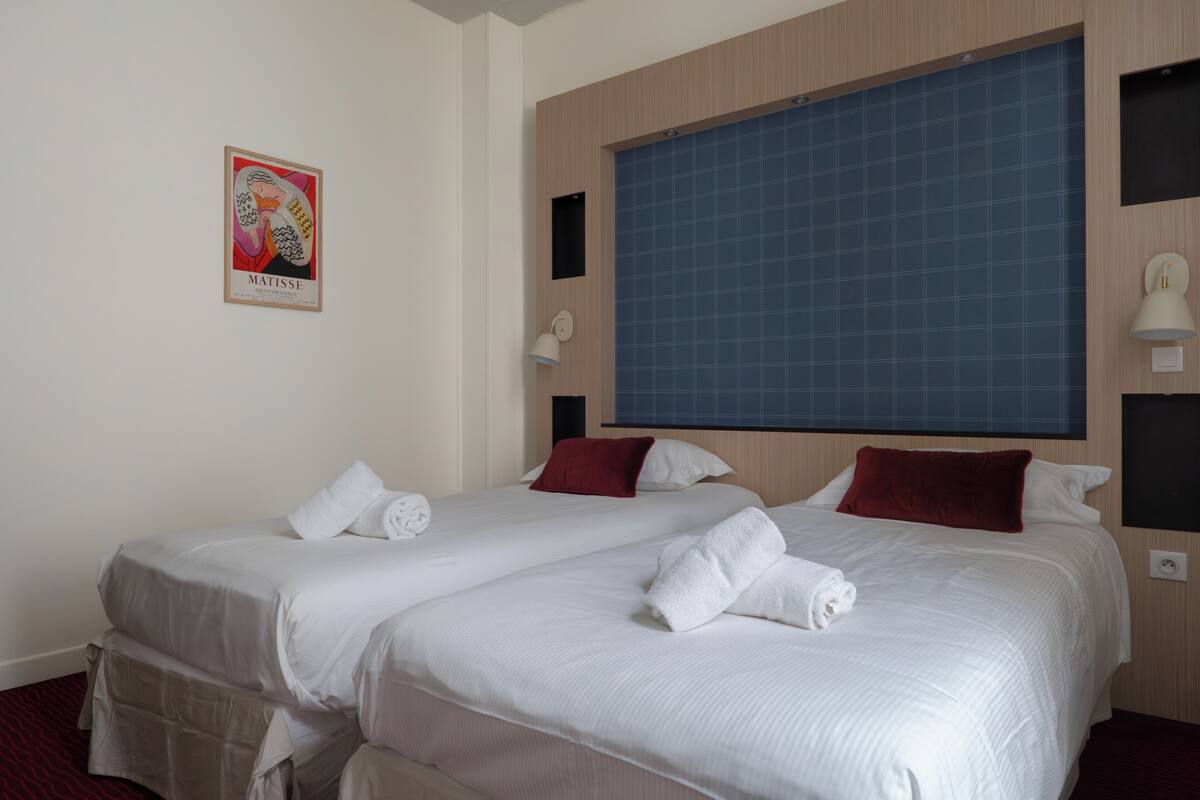 A hotel room with two beds, a large window, a wall-mounted lamp, and a framed artwork.
