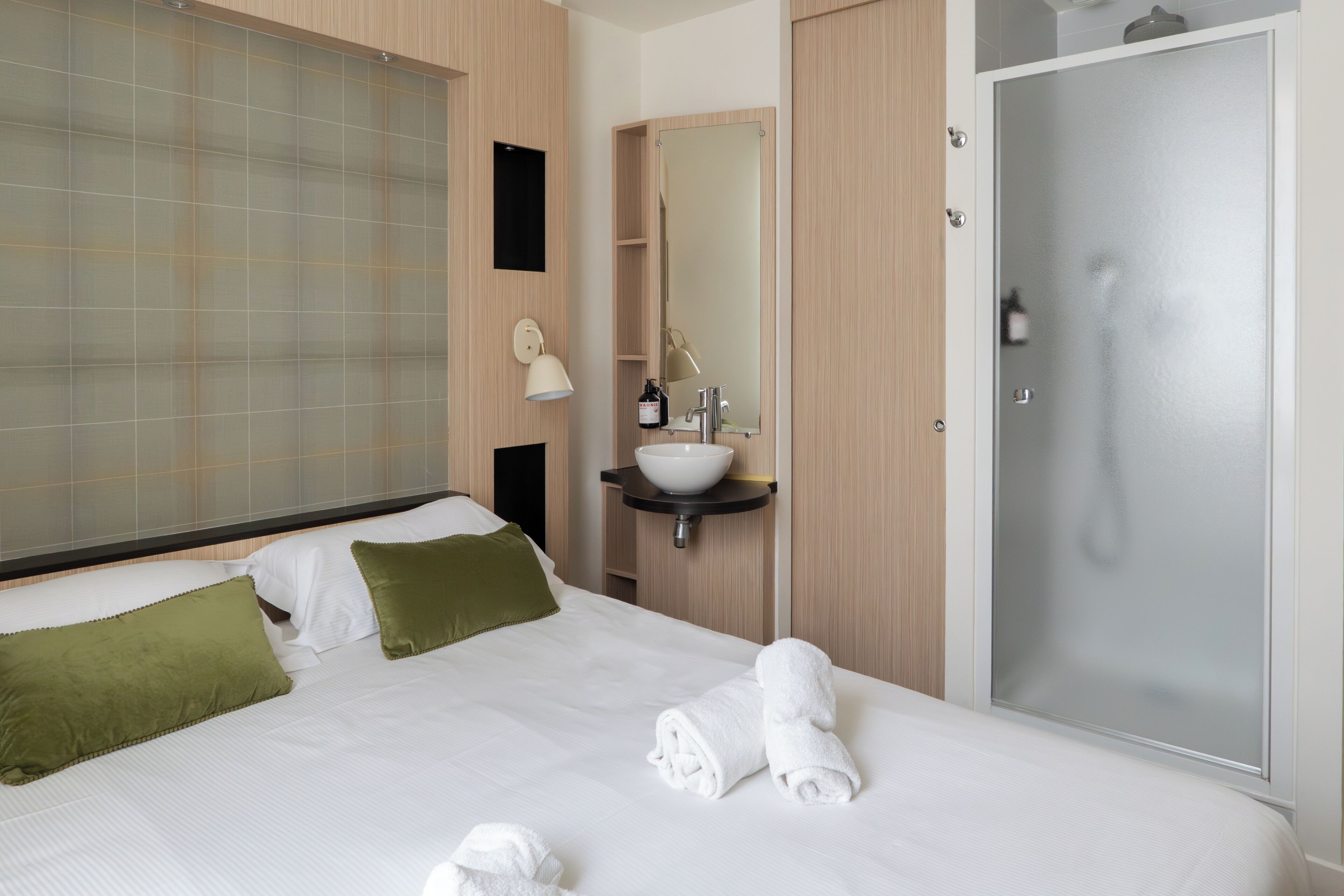 Una habitación de hotel con cama, mesita de noche con espejo, lavamanos y ducha.