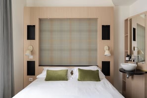 Ein Hotelzimmer mit Bett, großem Fenster mit Jalousien, Nachttisch mit Lampe und Badezimmer mit Waschbecken und Spiegel.