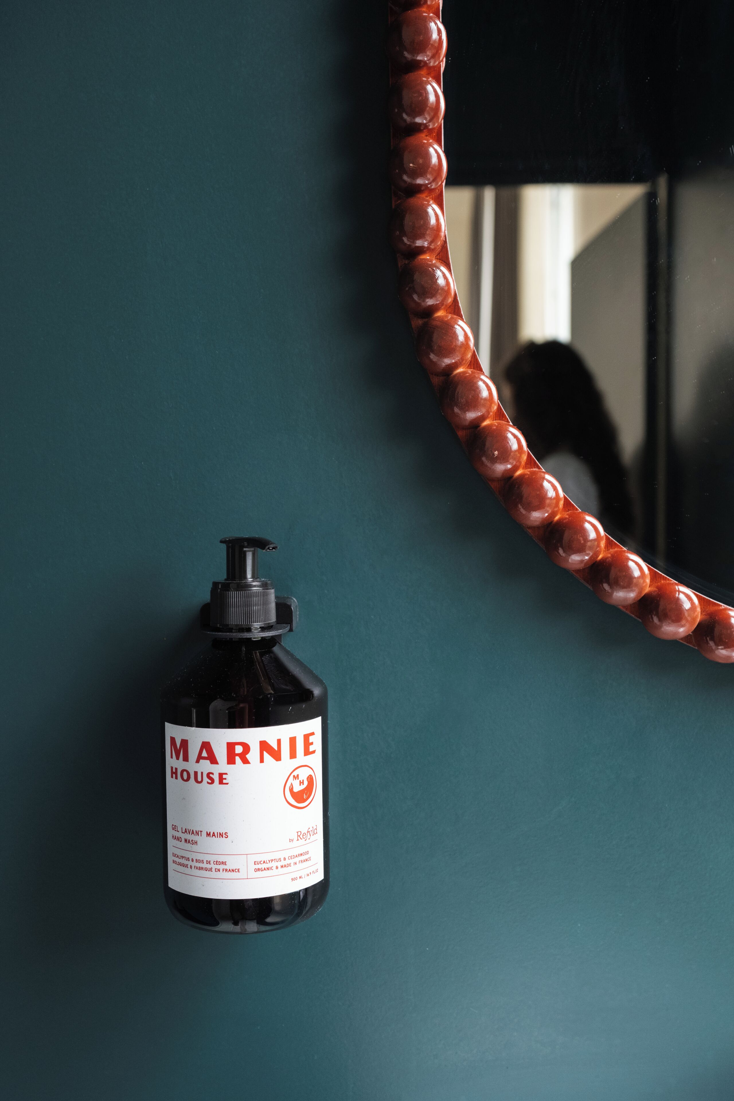 A bottle of Marnie House hand wash soap against a teal wall with a mirror reflecting a shadow.
