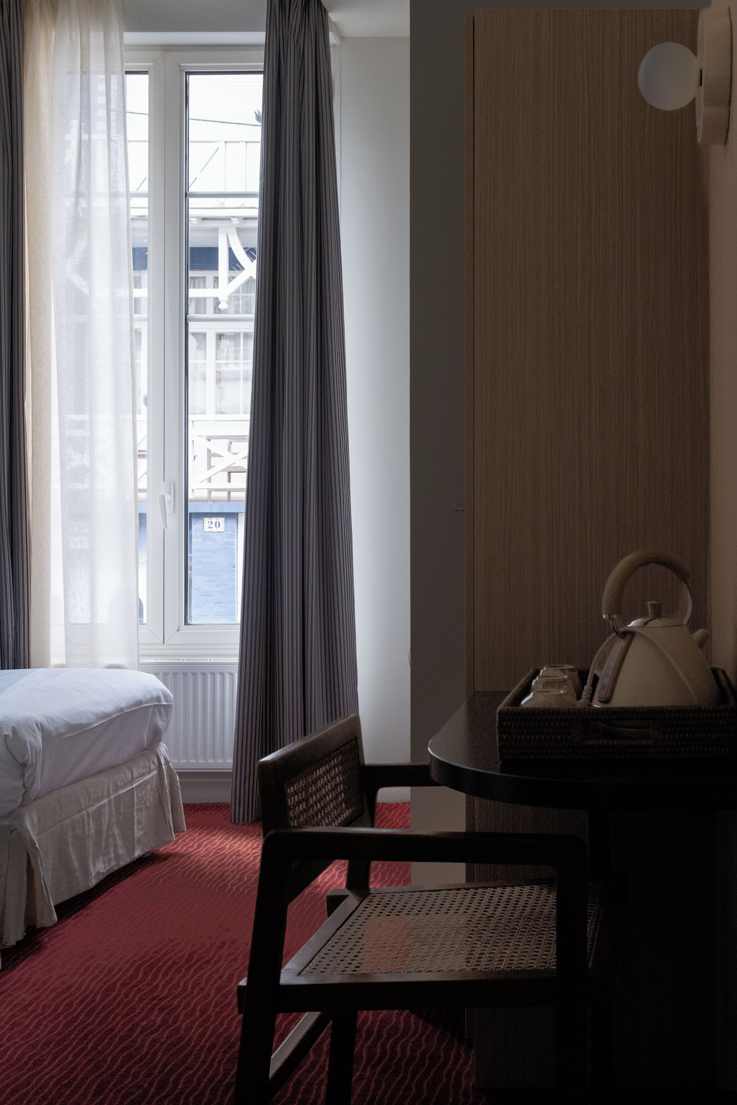 Habitación de hotel con cama, una silla, una mesa con hervidor y vista a un edificio a través de una ventana.
