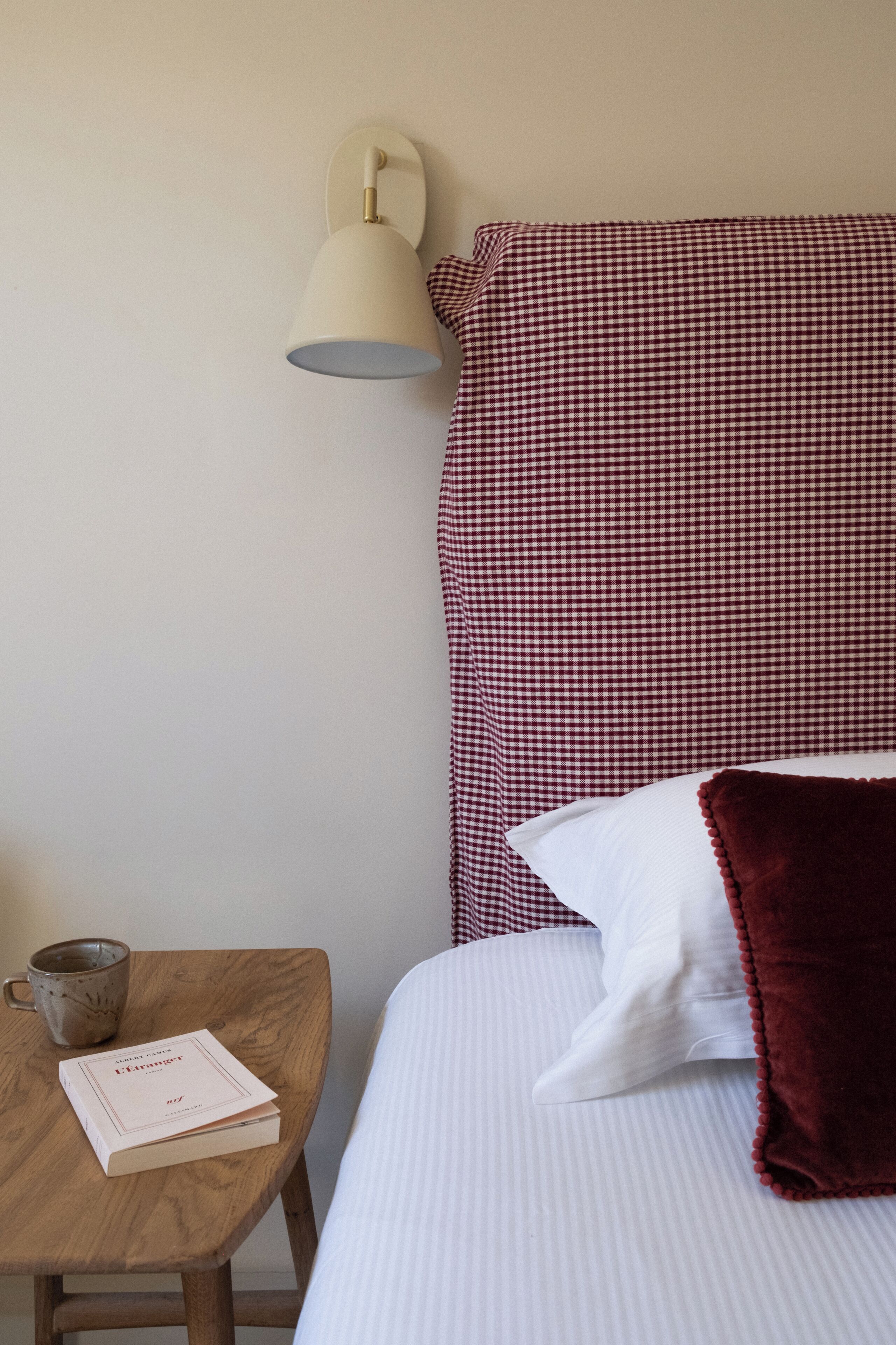 Un lit bien fait, avec un oreiller à carreaux et un oreiller blanc, une table de chevet sur laquelle se trouvent un livre et une tasse, et une lampe fixée au mur.