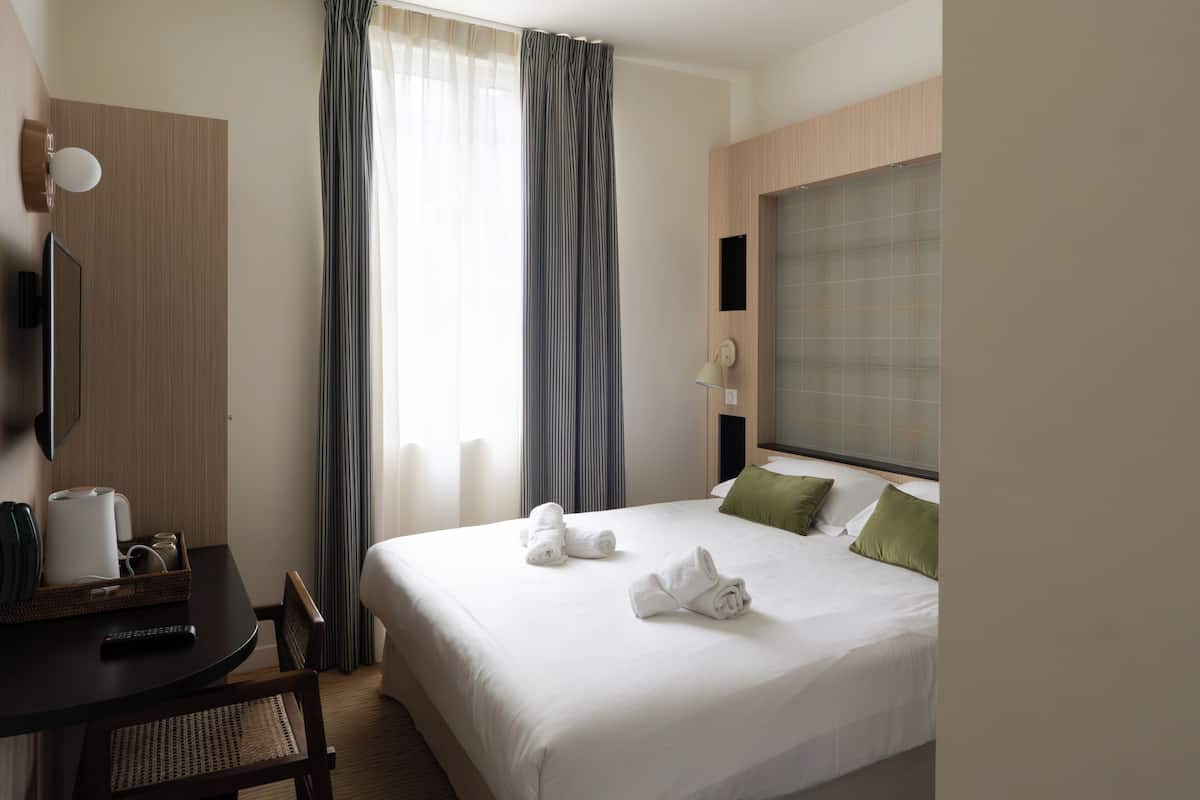 A hotel room with a bed, a desk with a chair, a television, and a window with curtains.