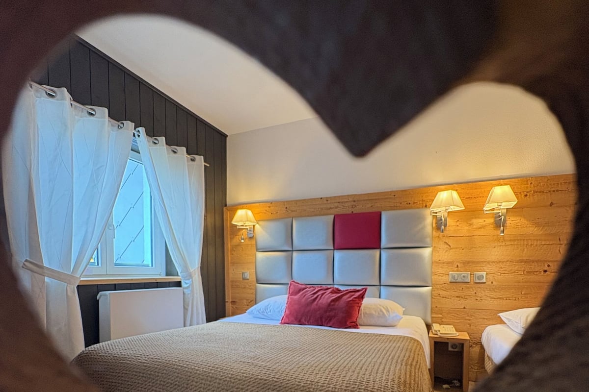 Una habitación de hotel con cama, mesita de noche, ventana con cortinas y una lámpara fijada a la pared.