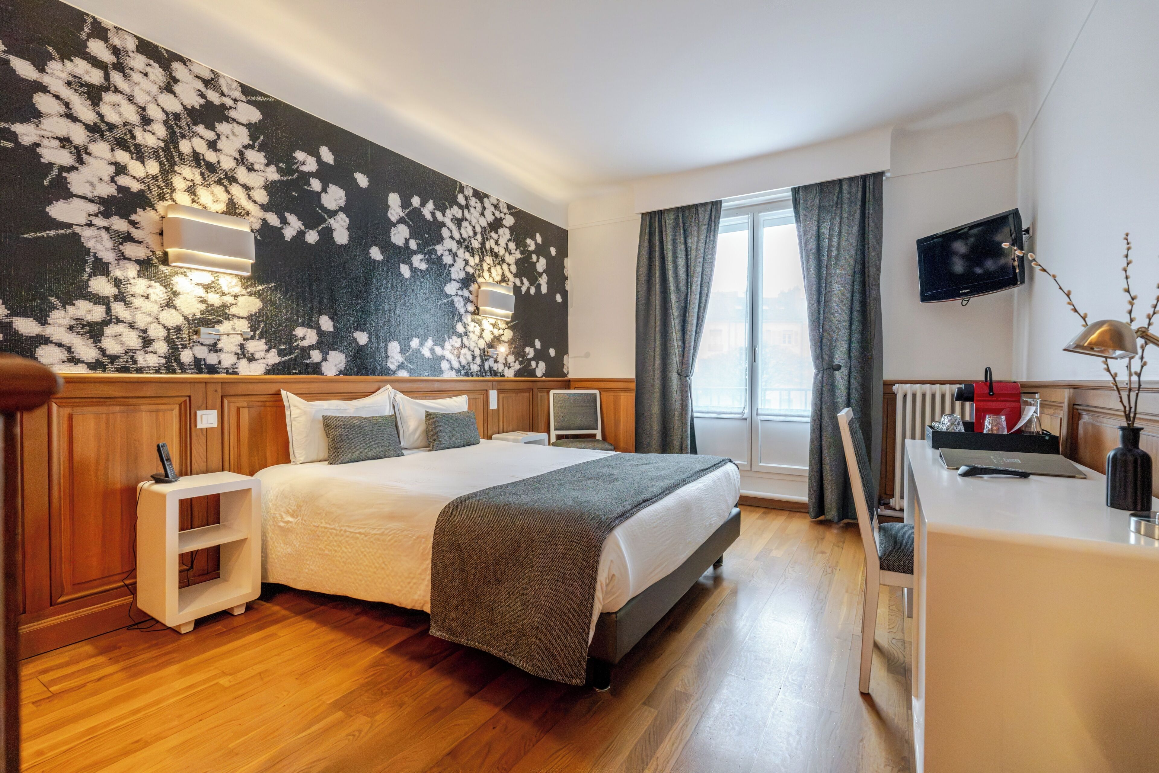 A hotel room with a bed, bedside tables, a desk, a TV, and a window with curtains.