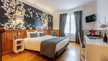 A hotel room with a bed, bedside tables, a desk, a TV, and a window with curtains.