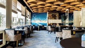 A restaurant with a high wooden ceiling, large windows, and a mural of a seascape. Tables are set with wine glasses and plates.