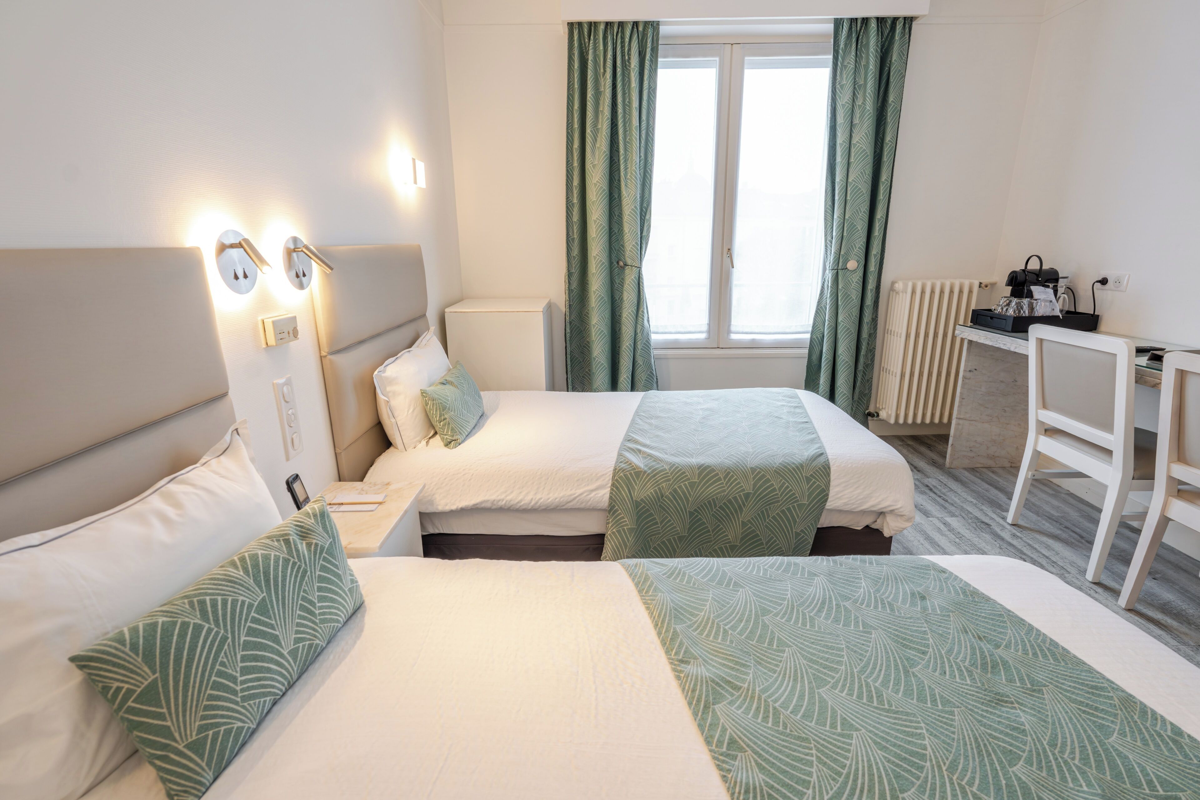 A hotel room with two beds, a desk, a radiator, and a window with curtains.
