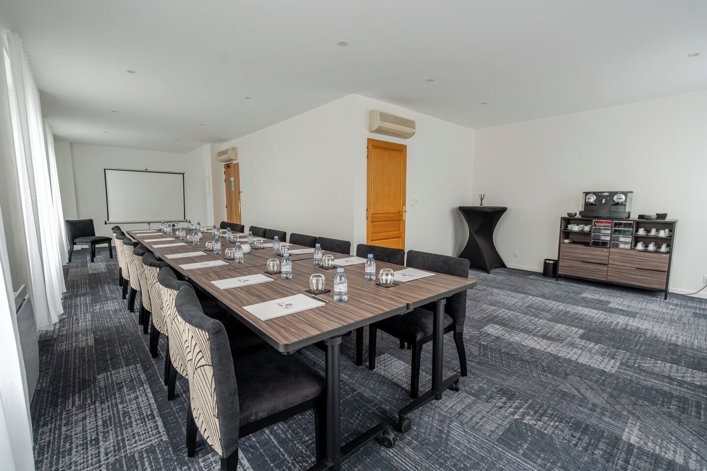 A conference room with a long table, chairs, water bottles, a projector screen, and a coffee station.