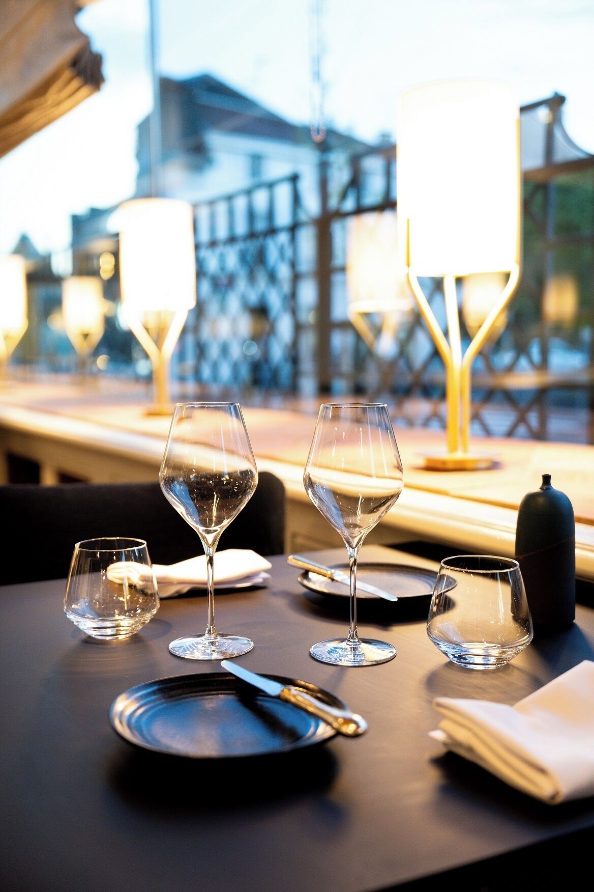 Una mesa con cuatro copas de vino, un cuchillo y un plato, frente a una vista de edificios y luces al fondo.