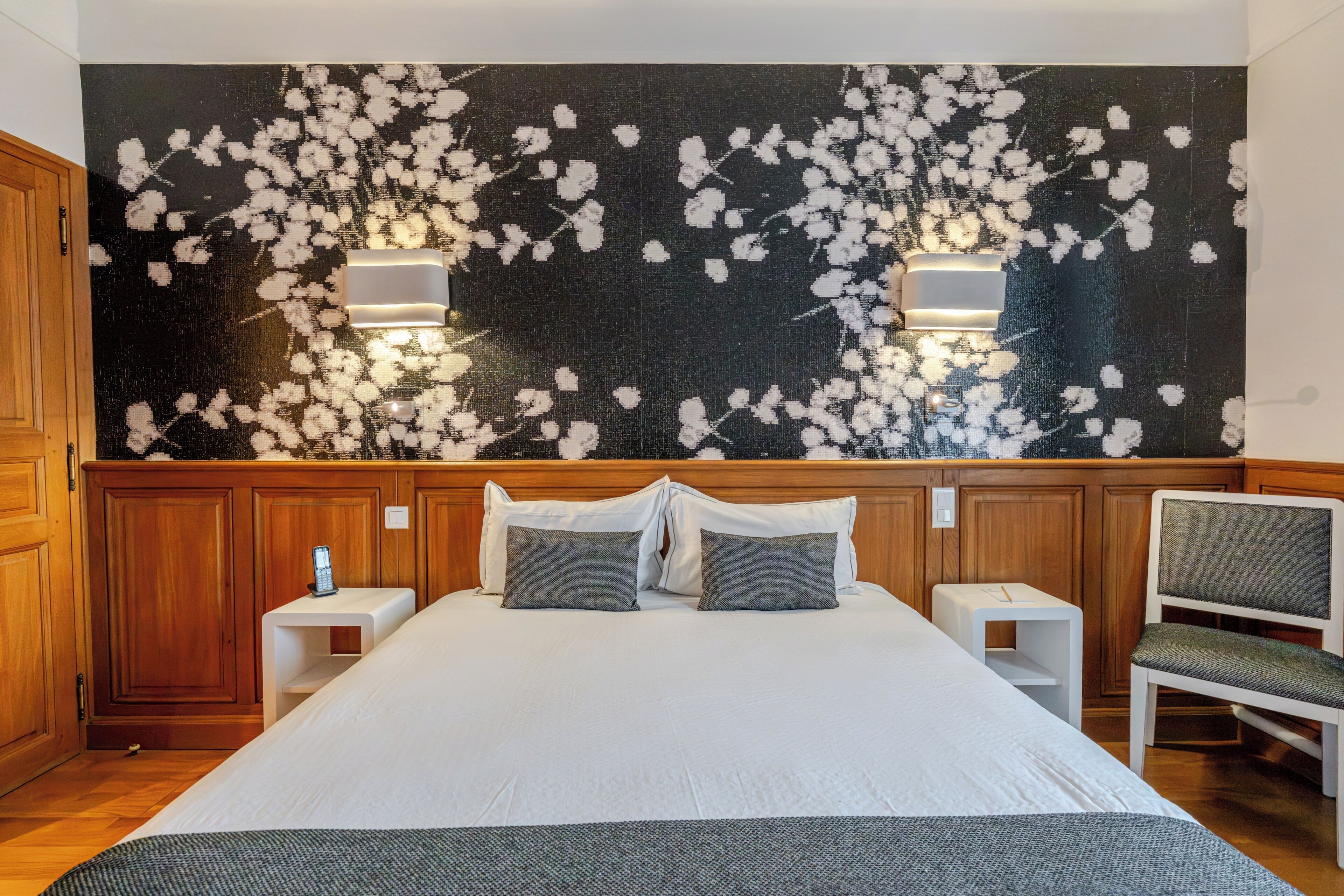 A bedroom with a large bed, two bedside tables, a chair, and a wall with a floral pattern.
