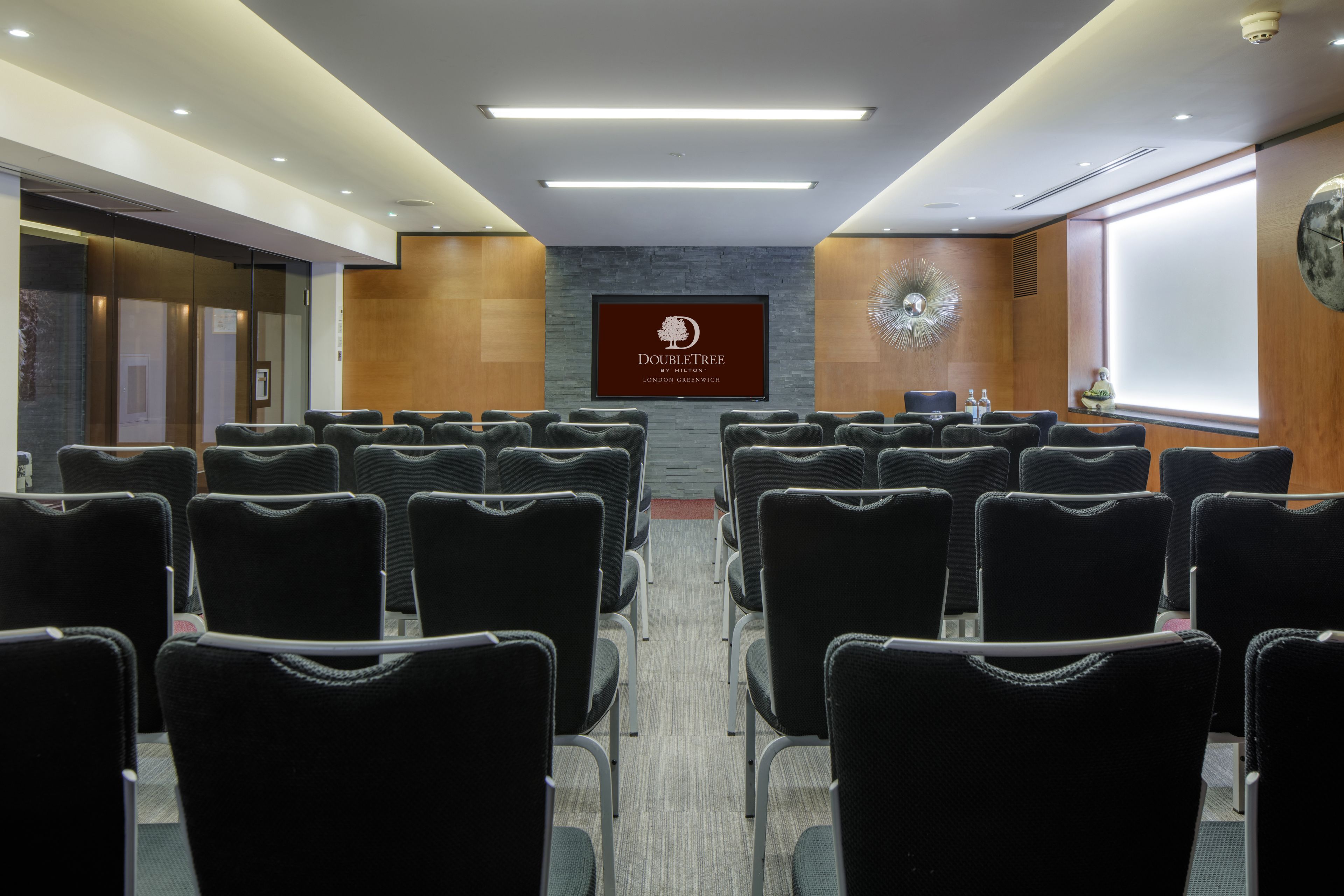 Un salón de conferencias con filas de sillas, una pantalla grande que muestra el logotipo de DoubleTree y un reloj en la pared.