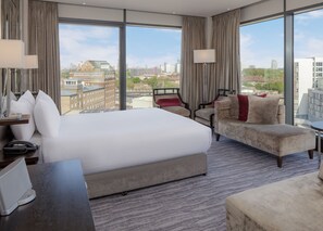 A hotel room with a large bed, a sofa, and a desk with a lamp. There are curtains and a view of the city. - DoubleTree by Hilton London - Greenwich (London)