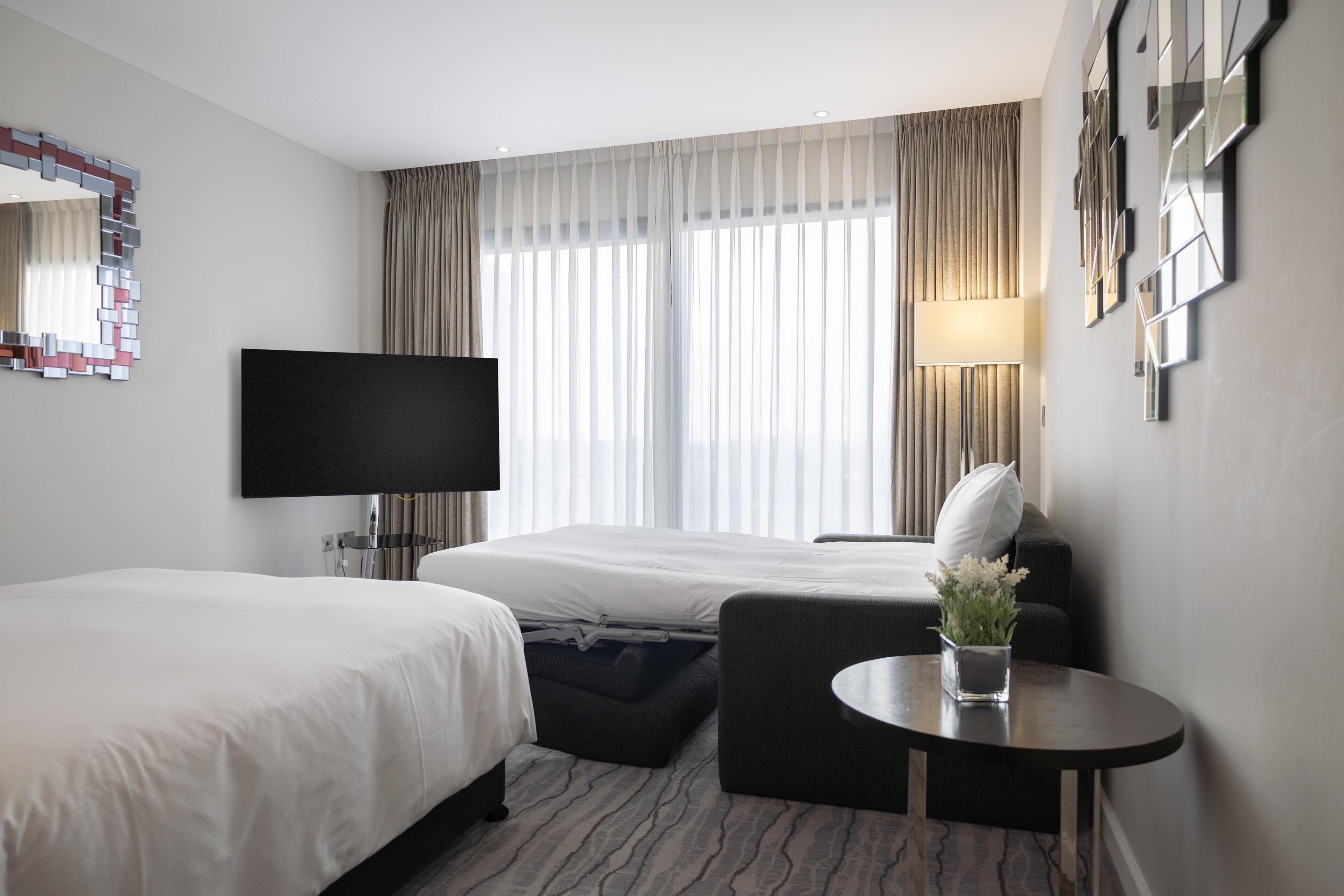 Quarto de hotel com duas camas, televisão, uma mesa pequena com um vaso e uma janela grande com cortinas.