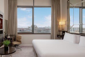 A hotel room with a bed, a desk, a chair, a TV, and a large window with a city view.