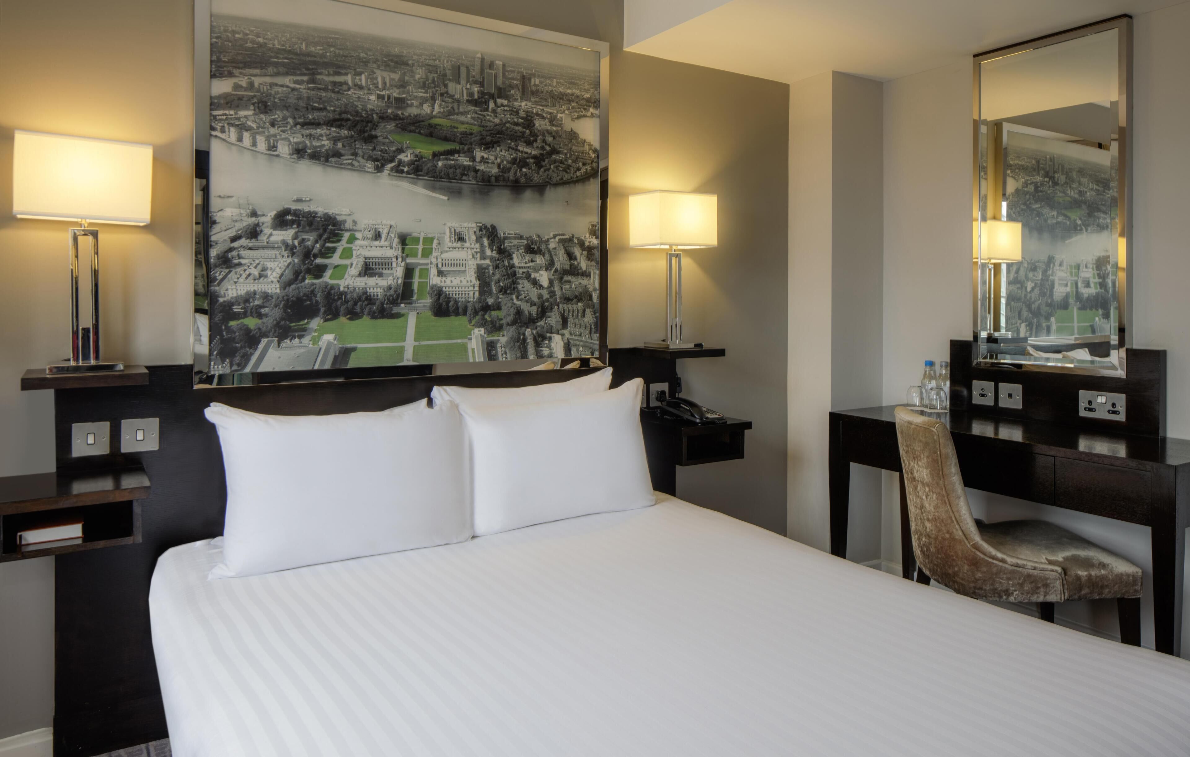 A hotel room with a large bed, two bedside tables with lamps, a chair, a large mirror, and a cityscape artwork on the wall.