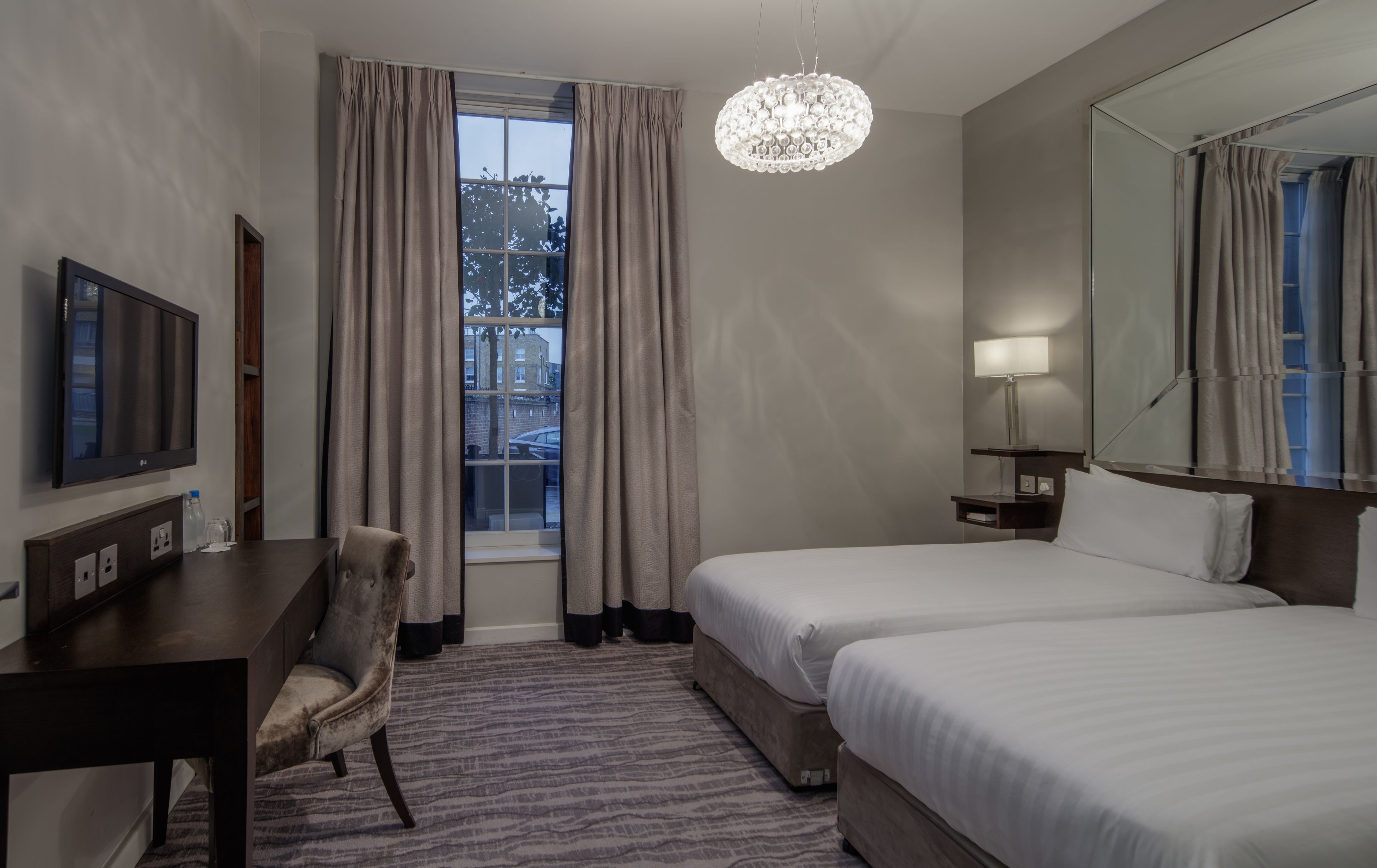 A hotel room with a bed, a desk, a chair, a TV, a lamp, and a large window with curtains.