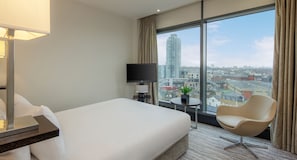 Ein Hotelzimmer mit einem großen Bett, einem Sessel, einem kleinen Tisch mit Pflanze, einem Fernseher und Blick auf die Stadt durch große Fenster.